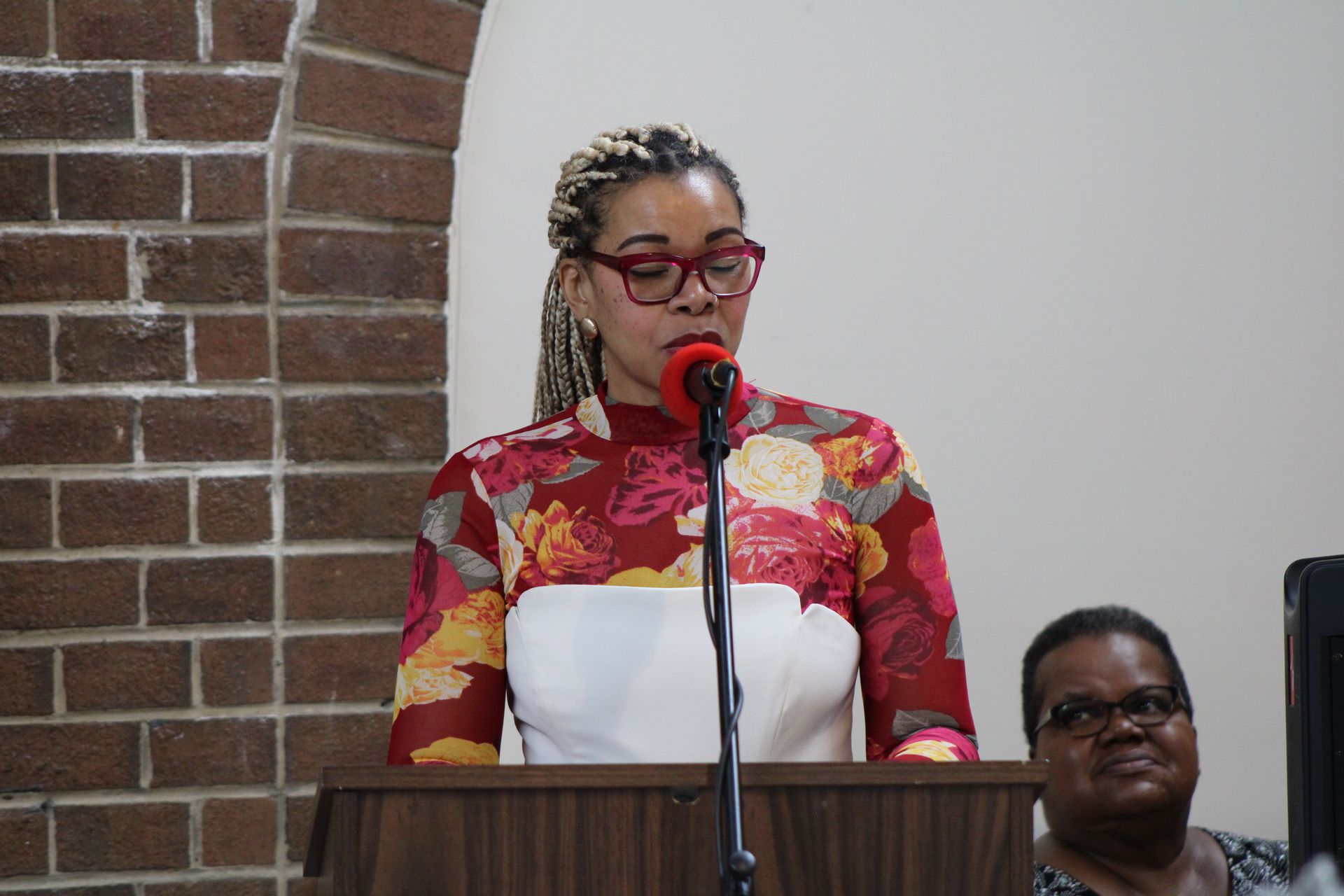 Woman speaking at a podium with a microphone, indoors near a brick wall.