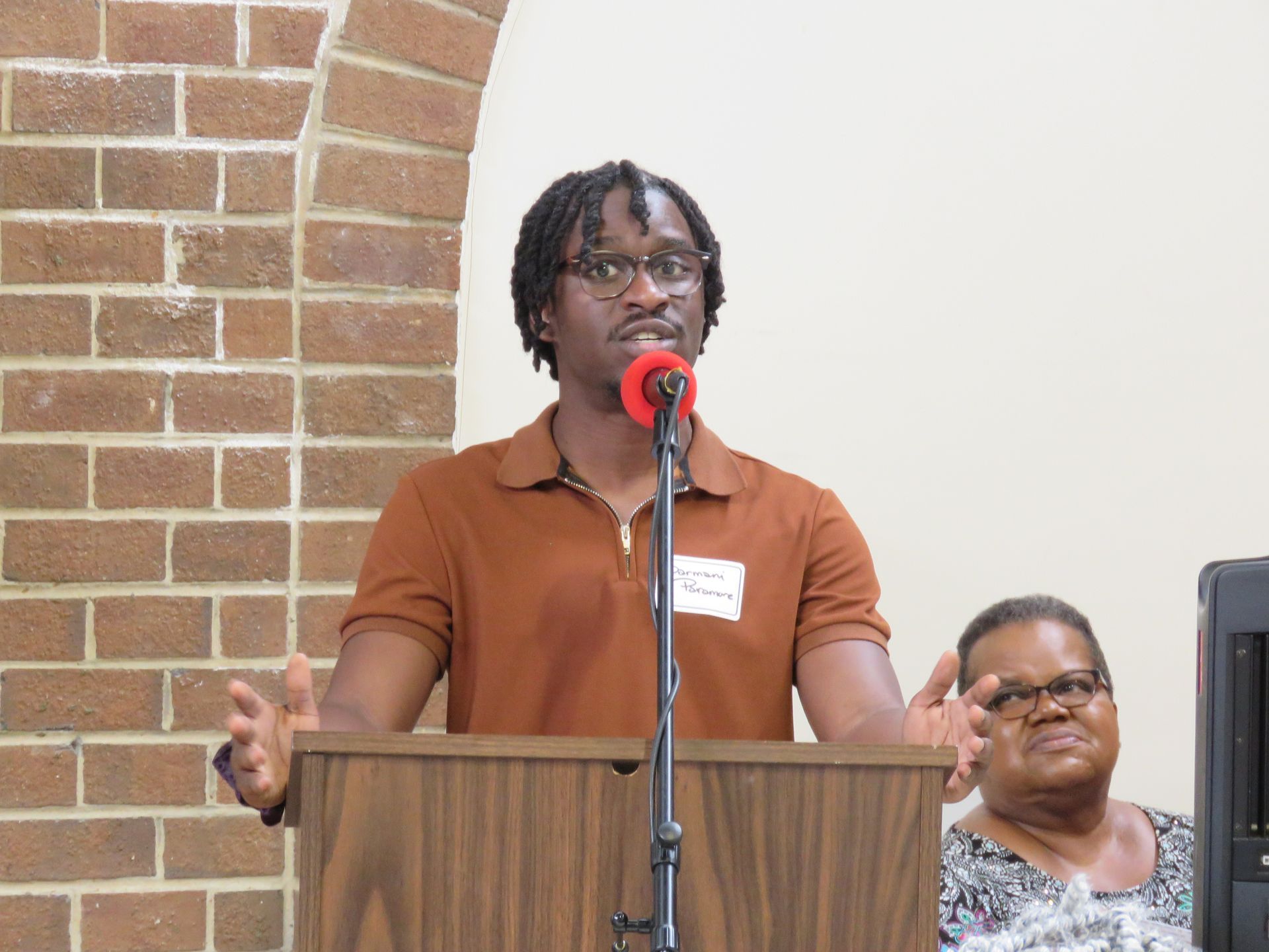 Man speaking at a podium with arms raised; brick wall and a woman seated behind.