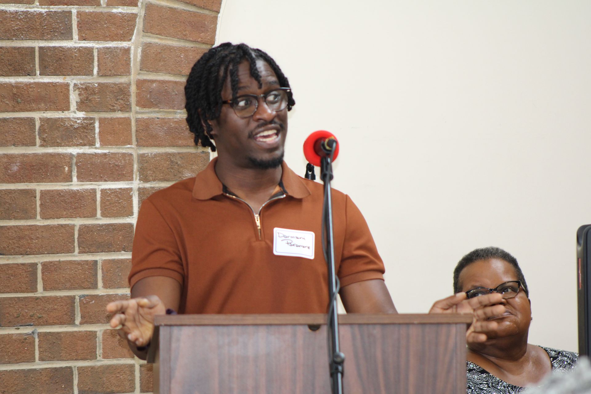 Man with dreadlocks speaking at a podium with a microphone, brick wall background.