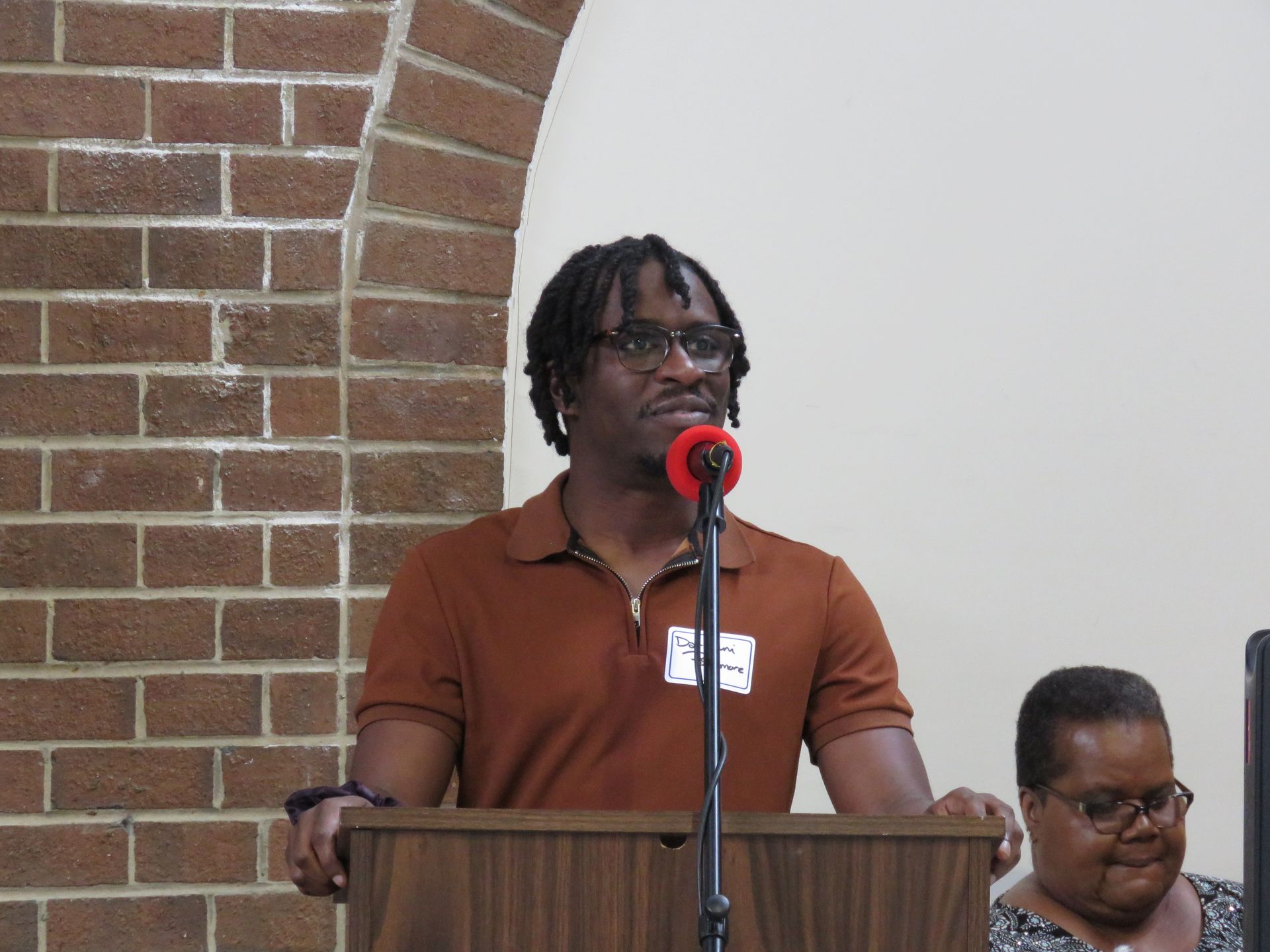 Man speaking at a podium with a microphone. Brick wall background.
