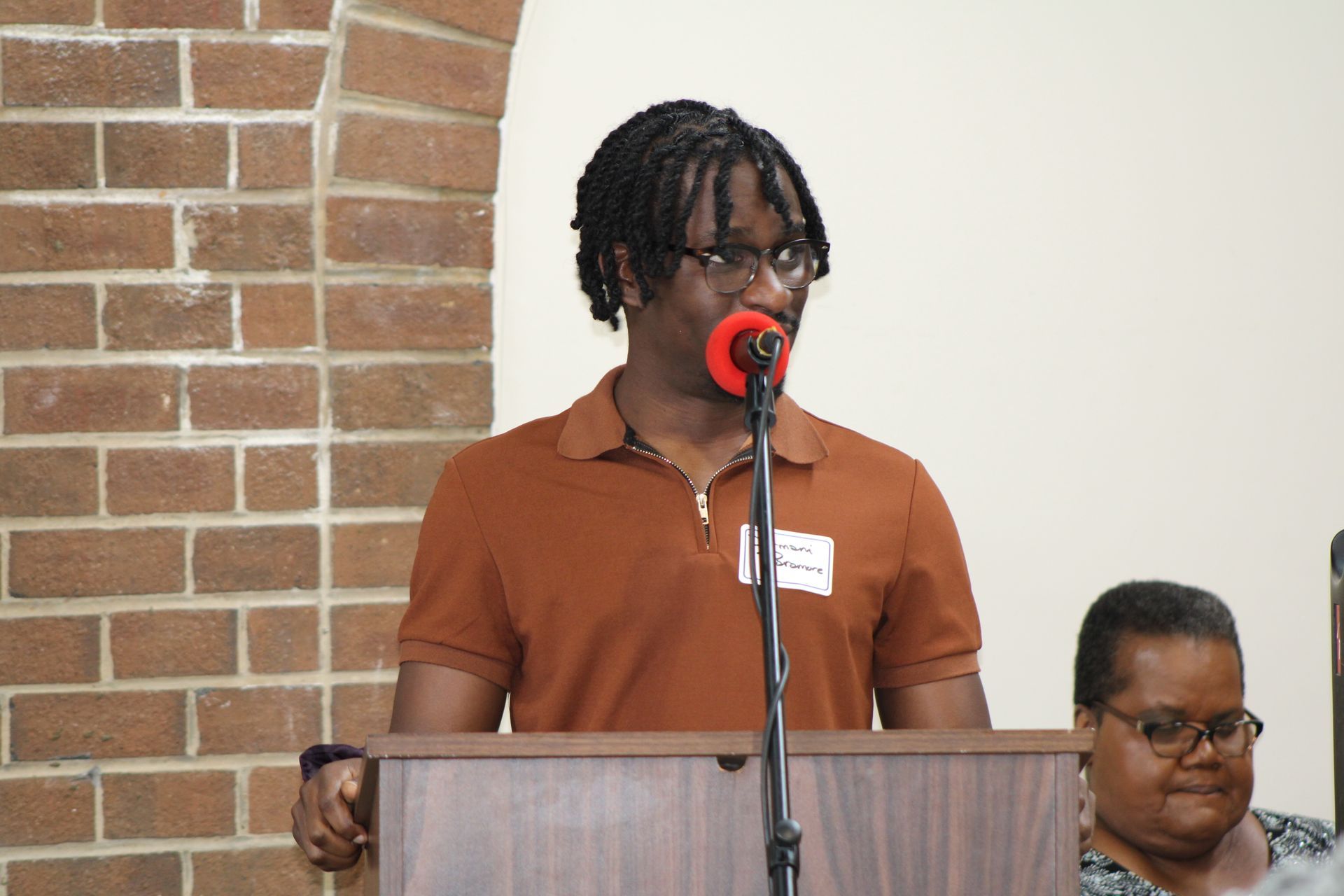 Person speaking at a podium with microphone in front of a brick wall, another person is sitting beside him.