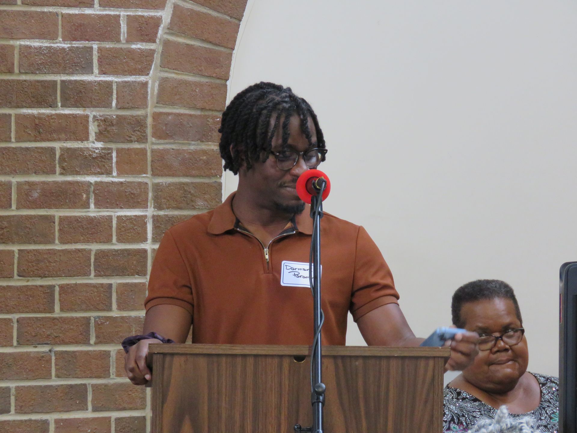 Man speaking at a podium, brown shirt, dreadlocks, red microphone, brick wall background.