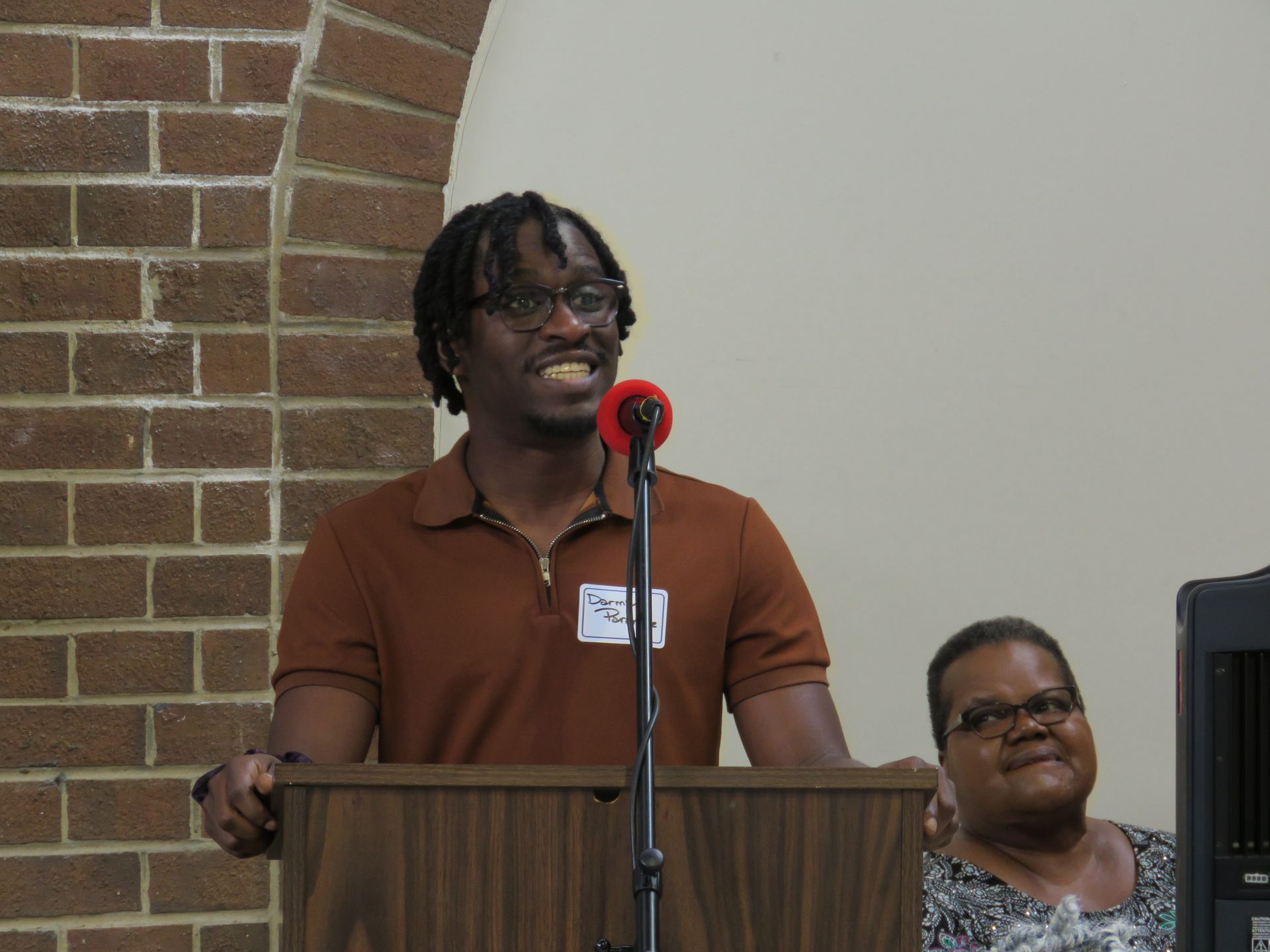Man speaking at a podium, smiling. Brick wall and woman seated visible.