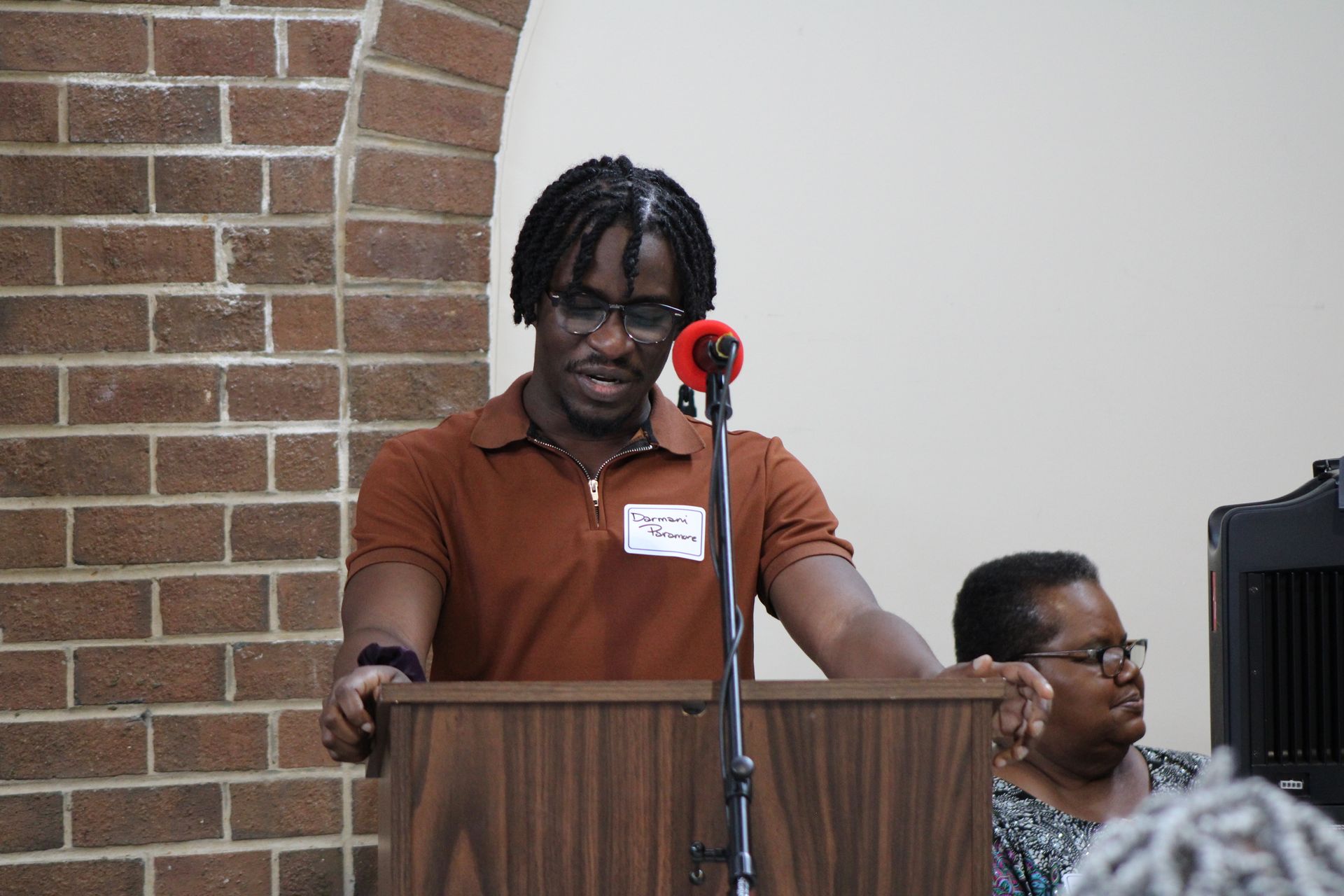 Man with dark hair speaks at a podium; brick wall and audience member in background.