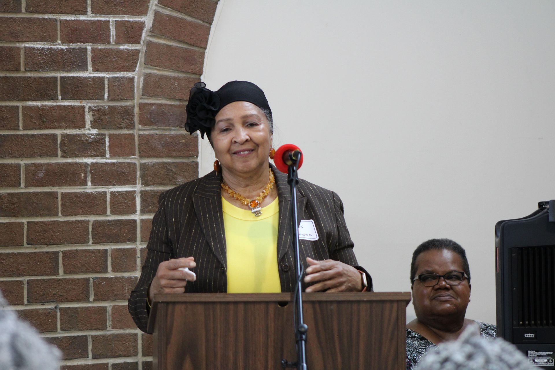 Woman speaking at a podium, wearing a blazer, with another woman seated nearby, brick wall background.