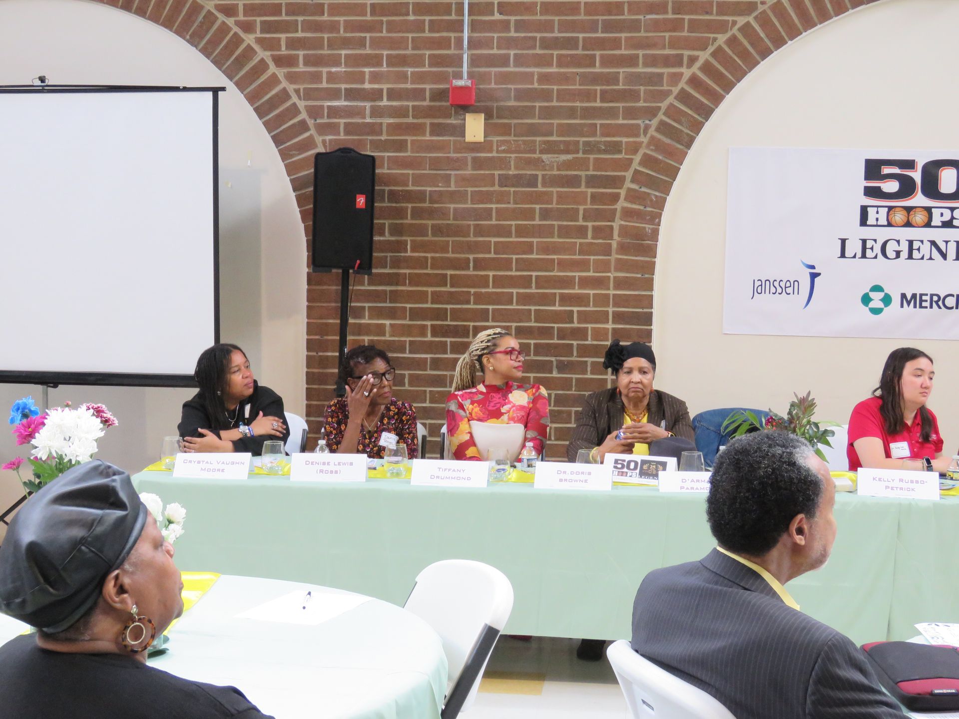 Panel of five speakers at a table with a white screen and an audience in a brick-walled room.