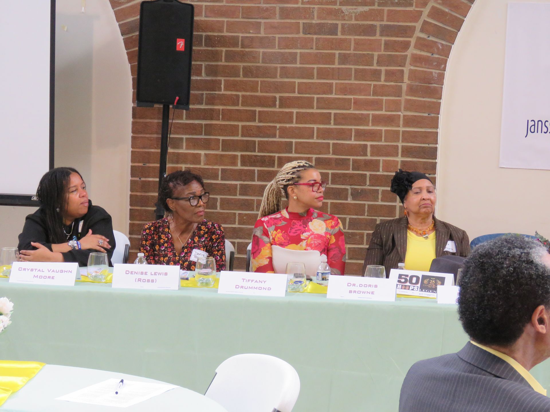 Panel of four women seated behind a table, speaking at an event in a brick-walled room.