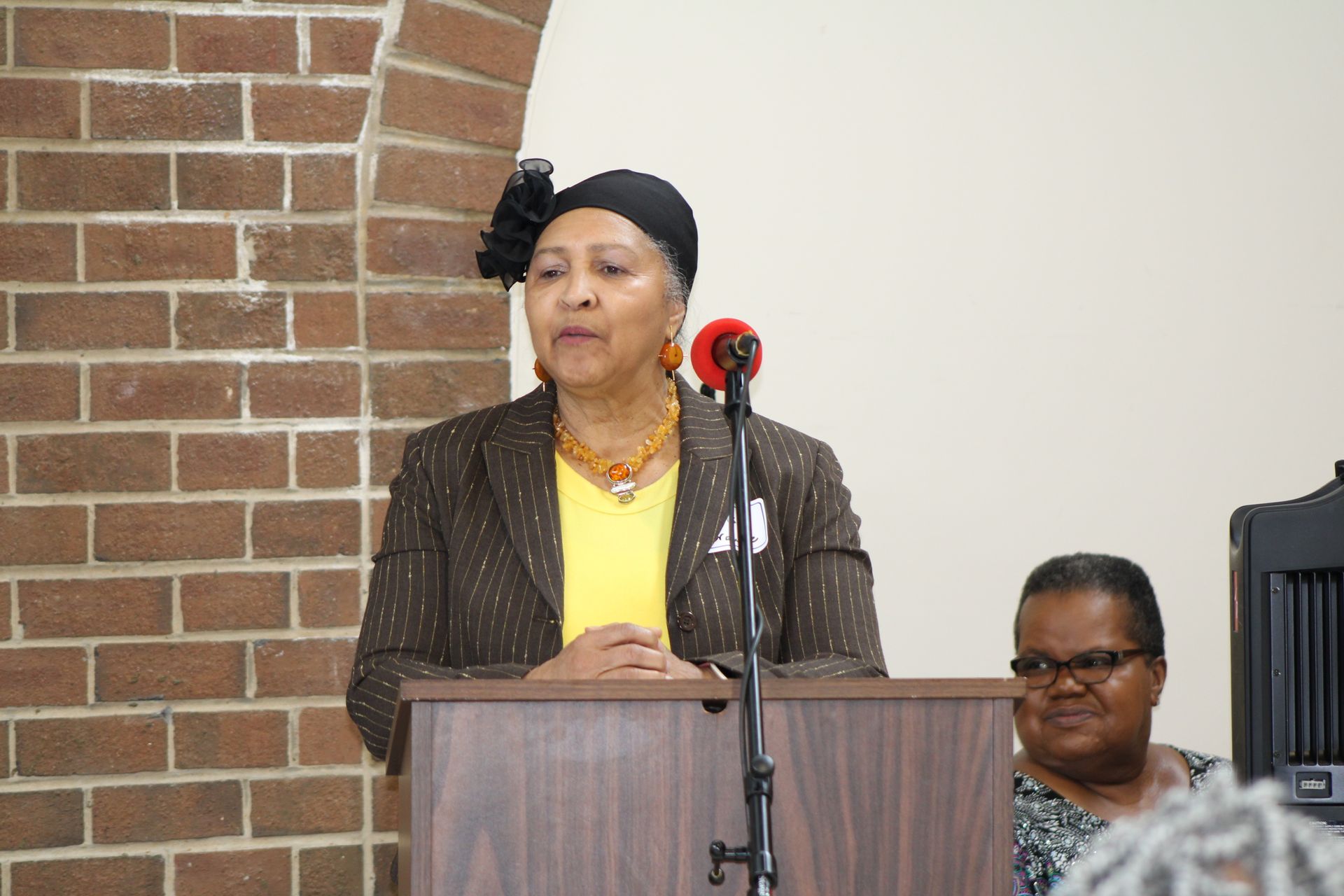 Woman speaking at a podium, brick wall background. Another woman sits nearby.