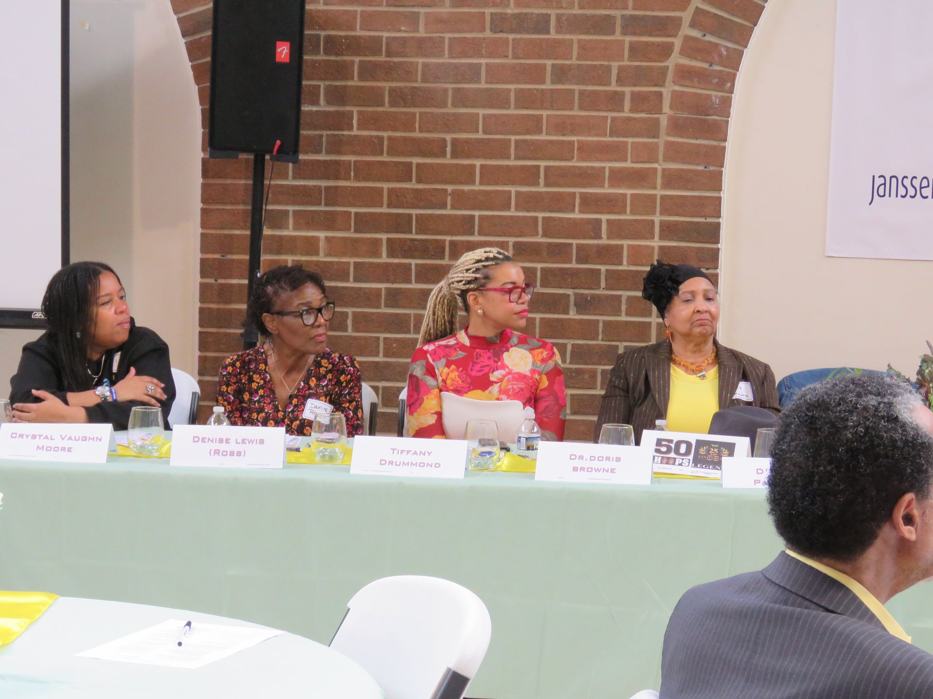 Panel of four women seated behind a table, speaking at an event in front of a brick wall.
