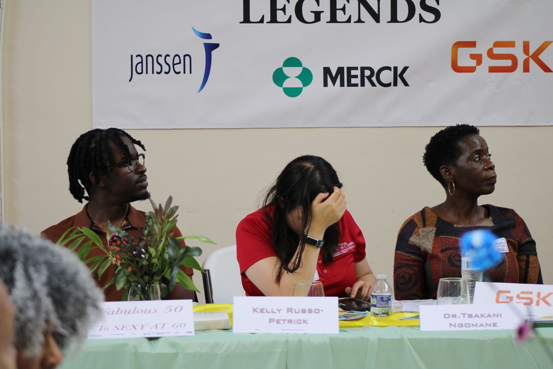 Three people seated at a table, one covering face with hand. Brands Janssen, Merck, and GSK on banner behind.