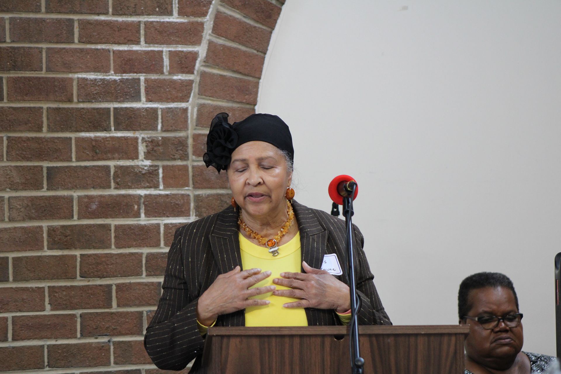 Woman speaking at a lectern, hands on chest, in front of brick wall.