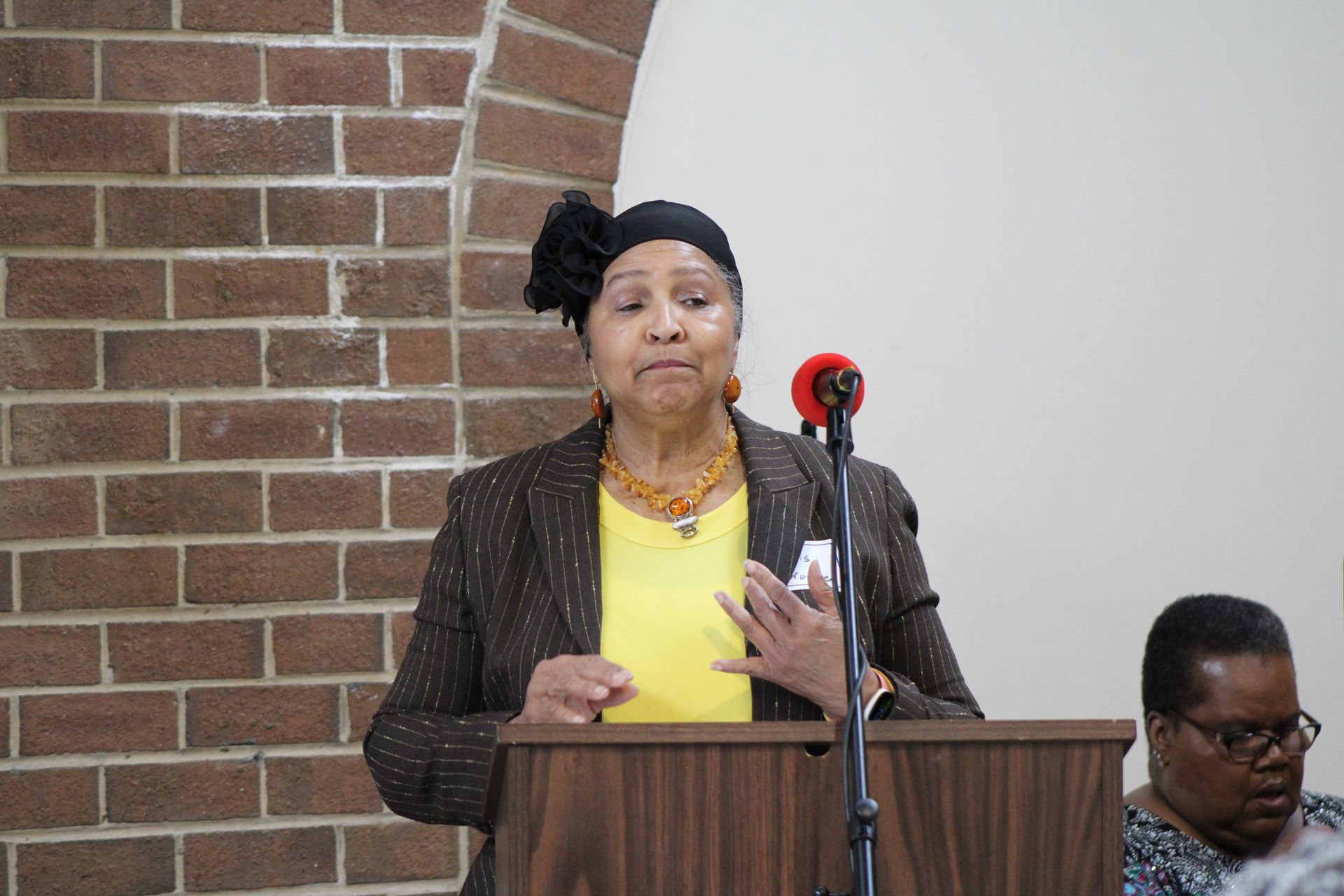 Woman speaking at a podium, near brick wall. She wears a blazer, yellow shirt, and a hat. Another woman sits to the right.