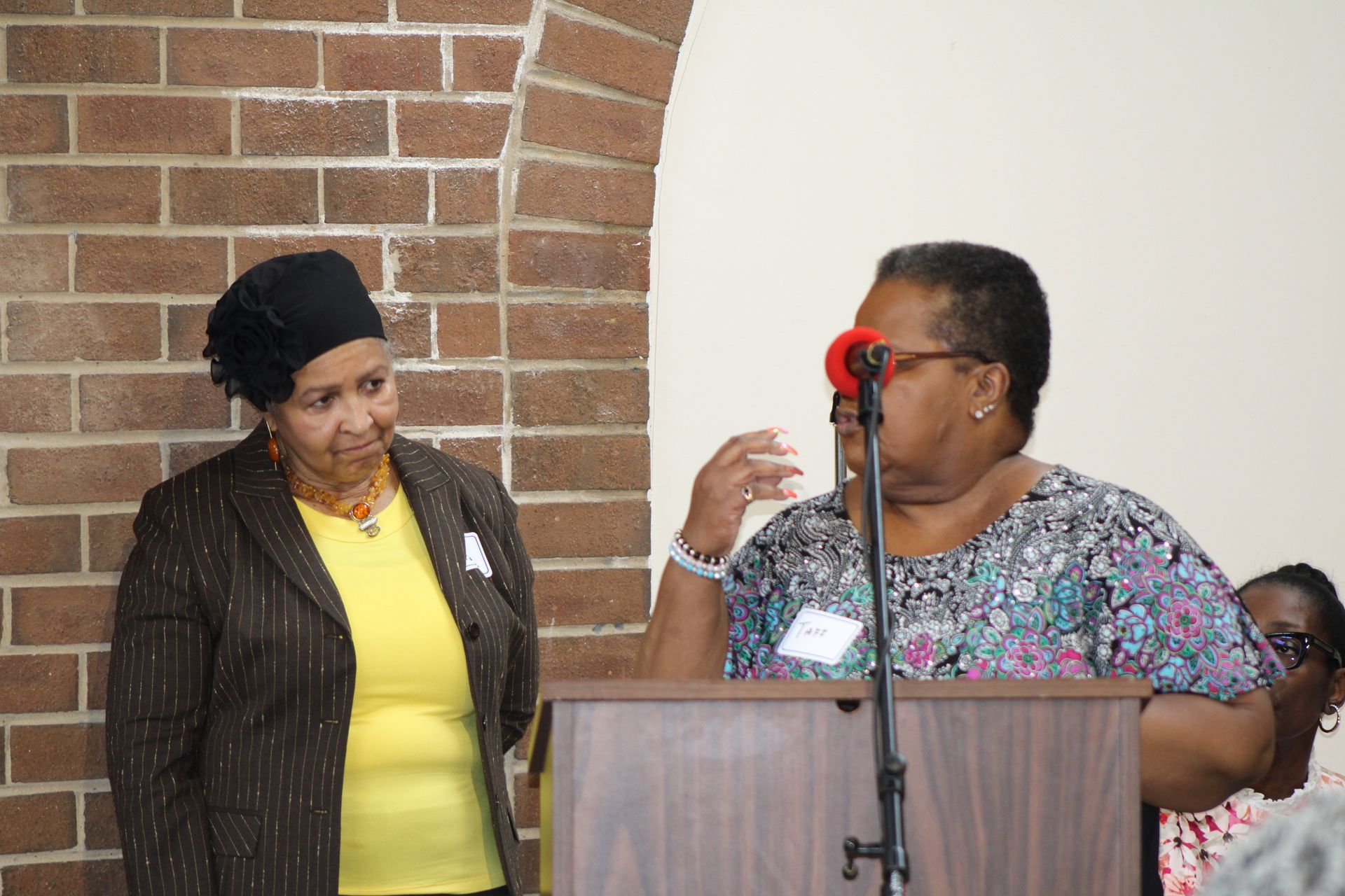 Two women at a podium. One speaks into a microphone, the other stands beside her. Brick wall in background.