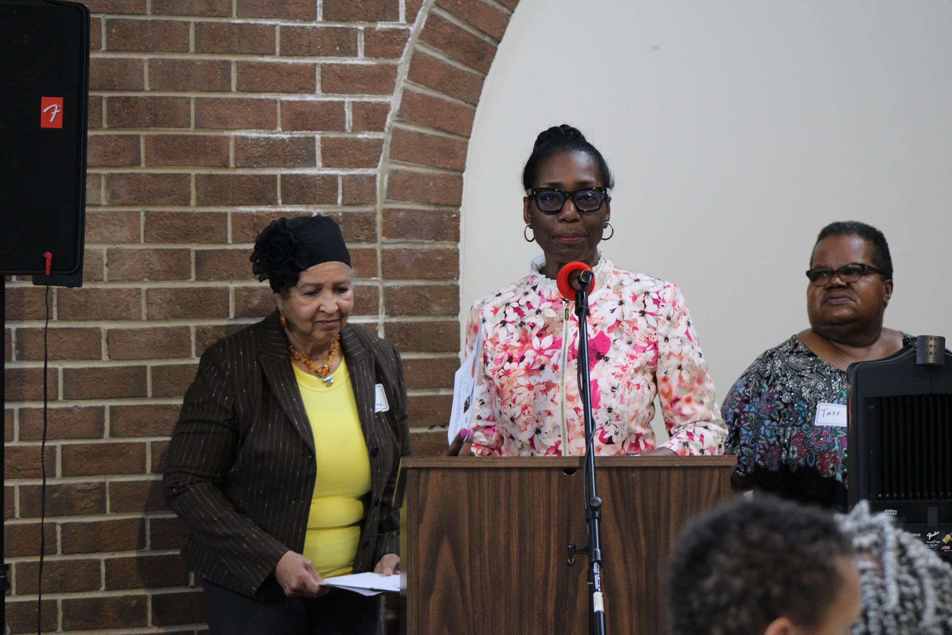 Woman speaking at podium; two women beside her in front of brick wall.