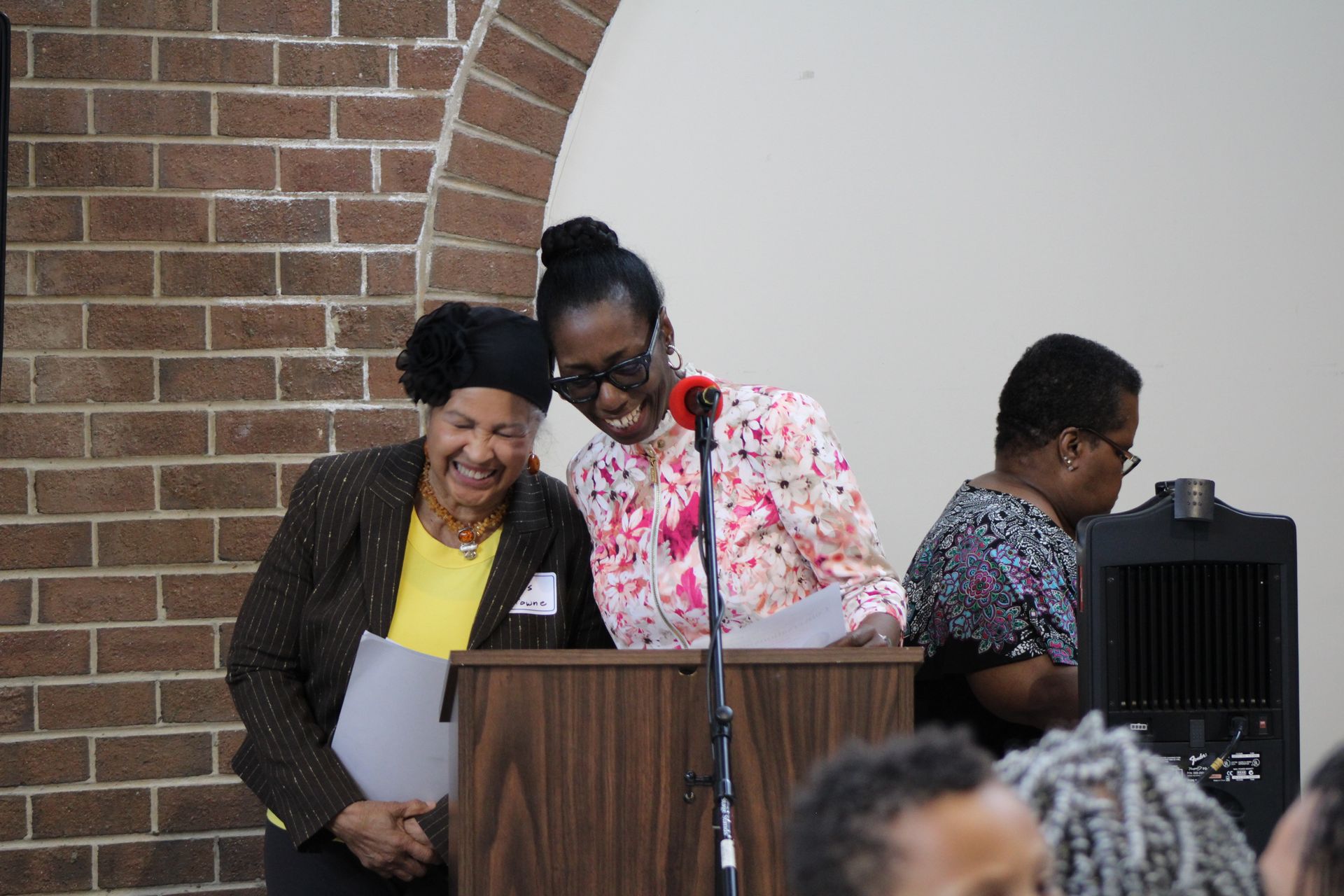 Two women laugh while reading at a podium. Brick wall and speaker in the background.