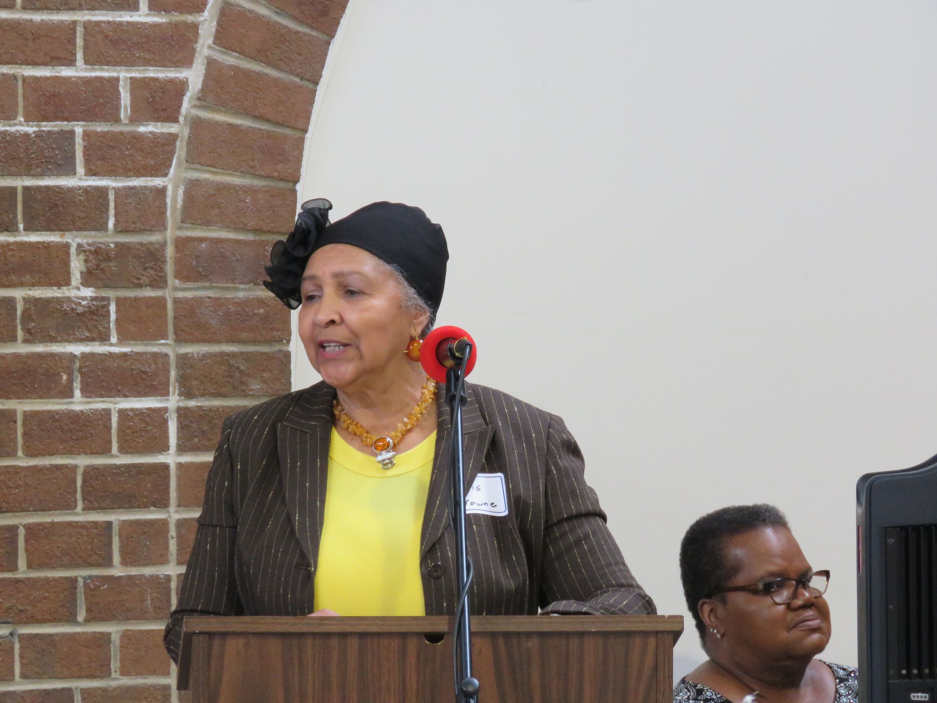 Woman speaking at a podium, wearing a dark hat and jacket. Brick arch and neutral wall in background.