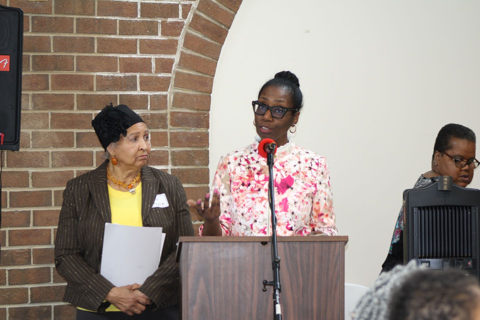 Woman speaks at a podium. Two women listen nearby, inside a room with a brick wall.