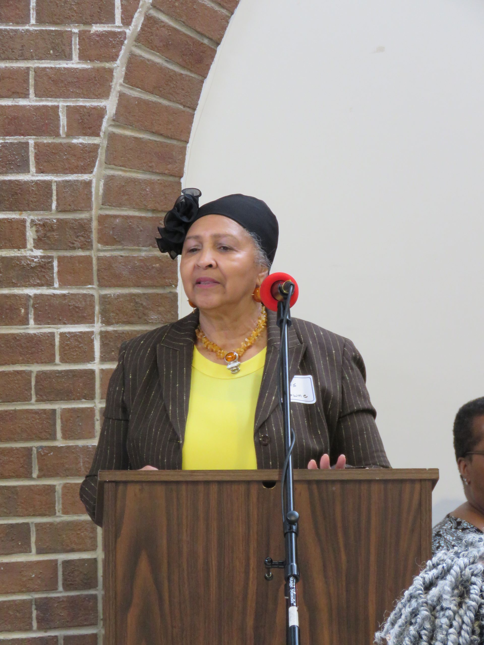 Woman speaking at a podium, wearing a black hat, brown jacket, and yellow shirt. Brick and white wall background.