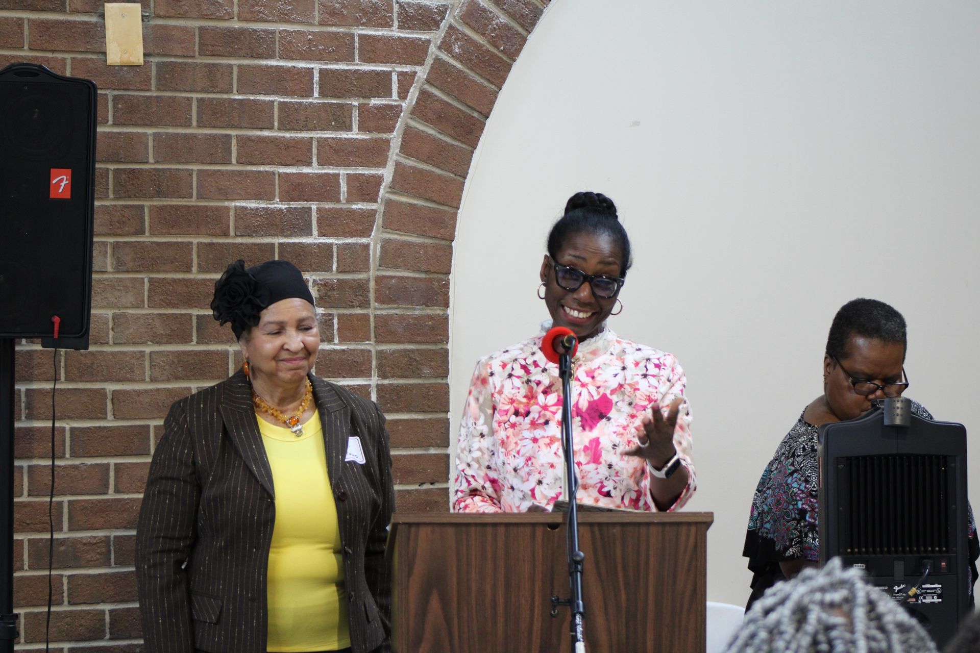 Three women at a podium in a brick-walled room. One speaks into a microphone, others stand on either side.
