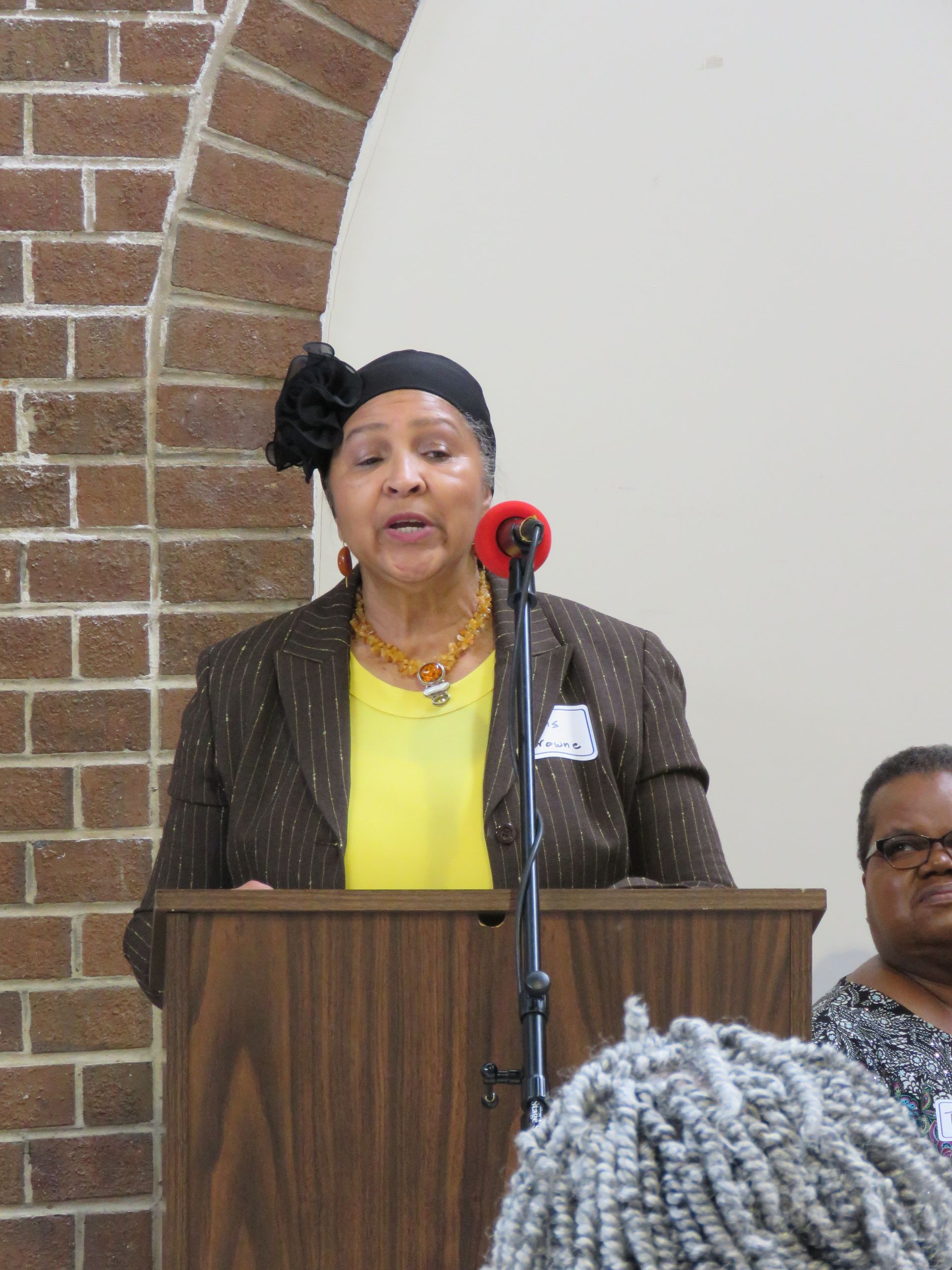 Woman speaking at a podium, wearing a blazer and yellow top. Brick archway in background.