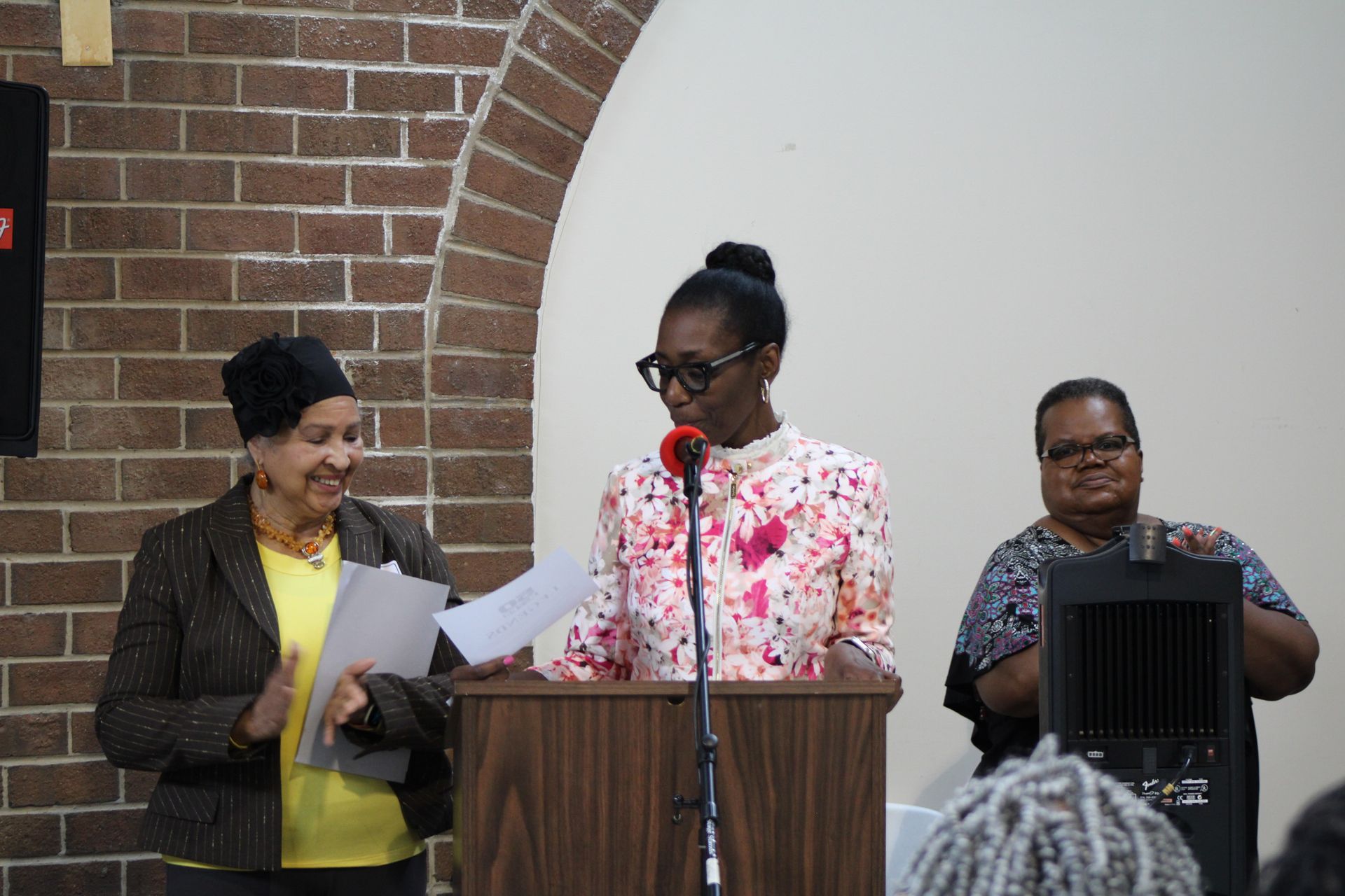 Three women at a podium, one speaking with papers. Two others stand beside, one with a speaker. Brick wall background.