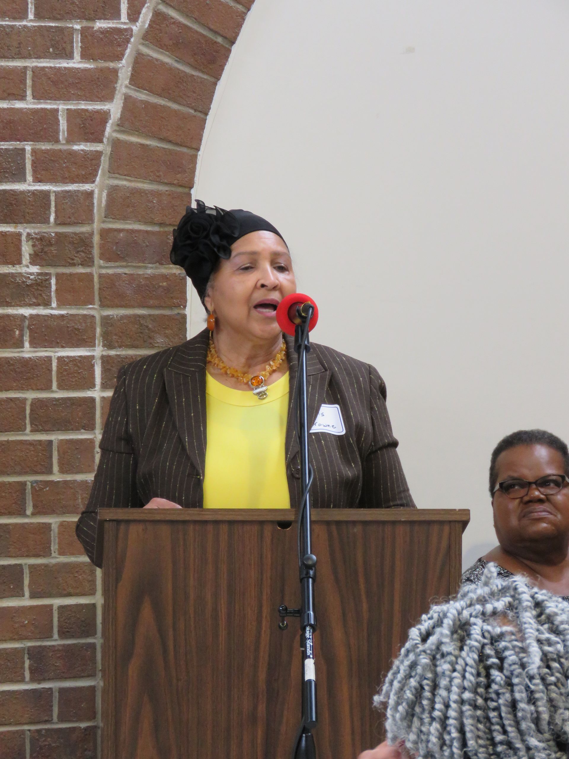 Woman speaking at a lectern, wearing a blazer, yellow shirt, and a hat. Red microphone. Brick wall in background.