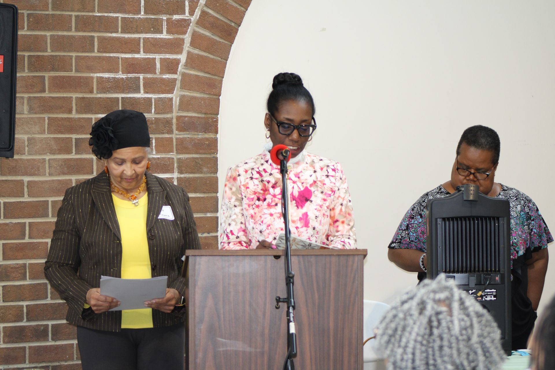 Three women at a podium, one speaking with a microphone, in front of a brick and white wall.