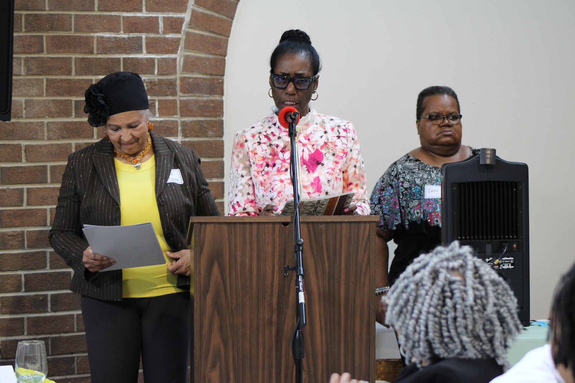 Three women at a podium, one speaking with a microphone. Others hold papers, brick wall in background.