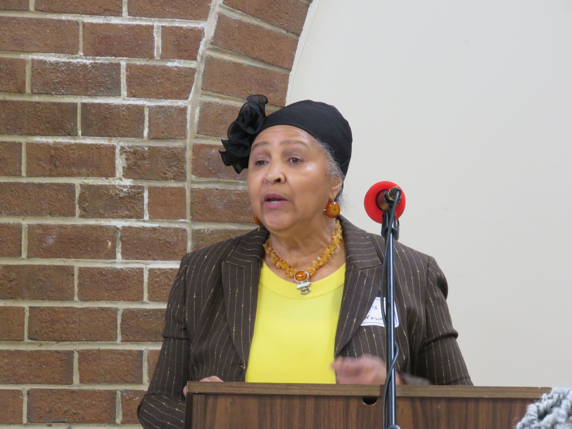 Woman speaking at a podium, wearing a dark head covering, yellow shirt, brown jacket, and necklace. Brick wall background.