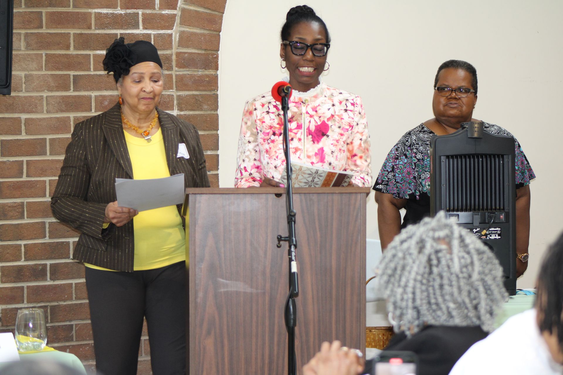 Three women at podium, speaking. Woman in center with glasses, flower print top. Others hold papers, speak at an event.
