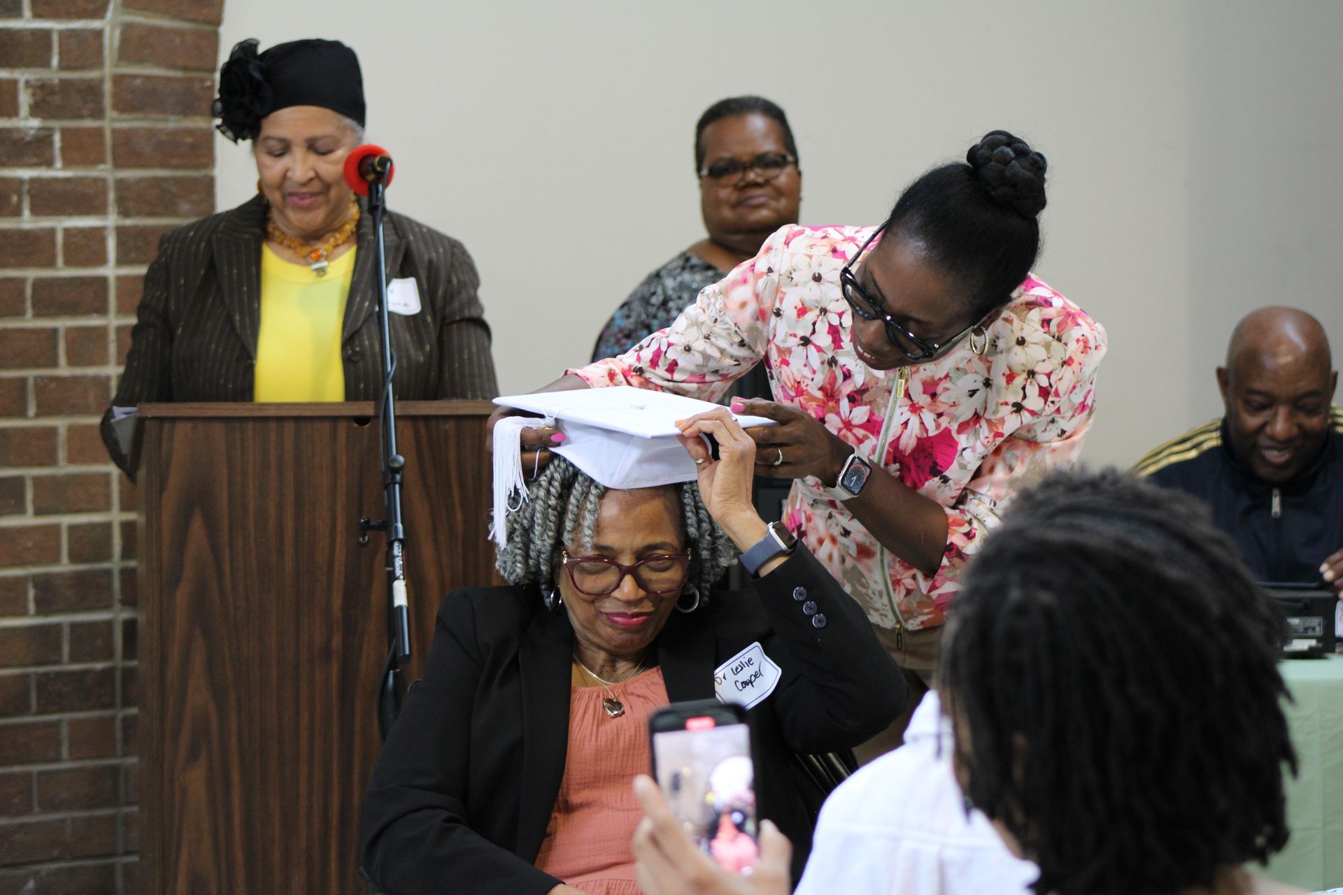 Woman in floral shirt places a mortarboard on another woman. Other people stand nearby in a room.