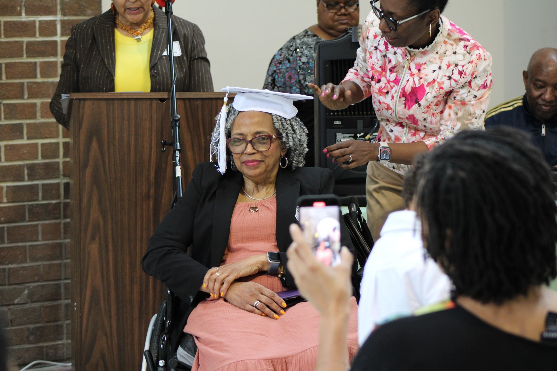 Woman in wheelchair wearing a graduation cap, with help adjusting it. People in background.