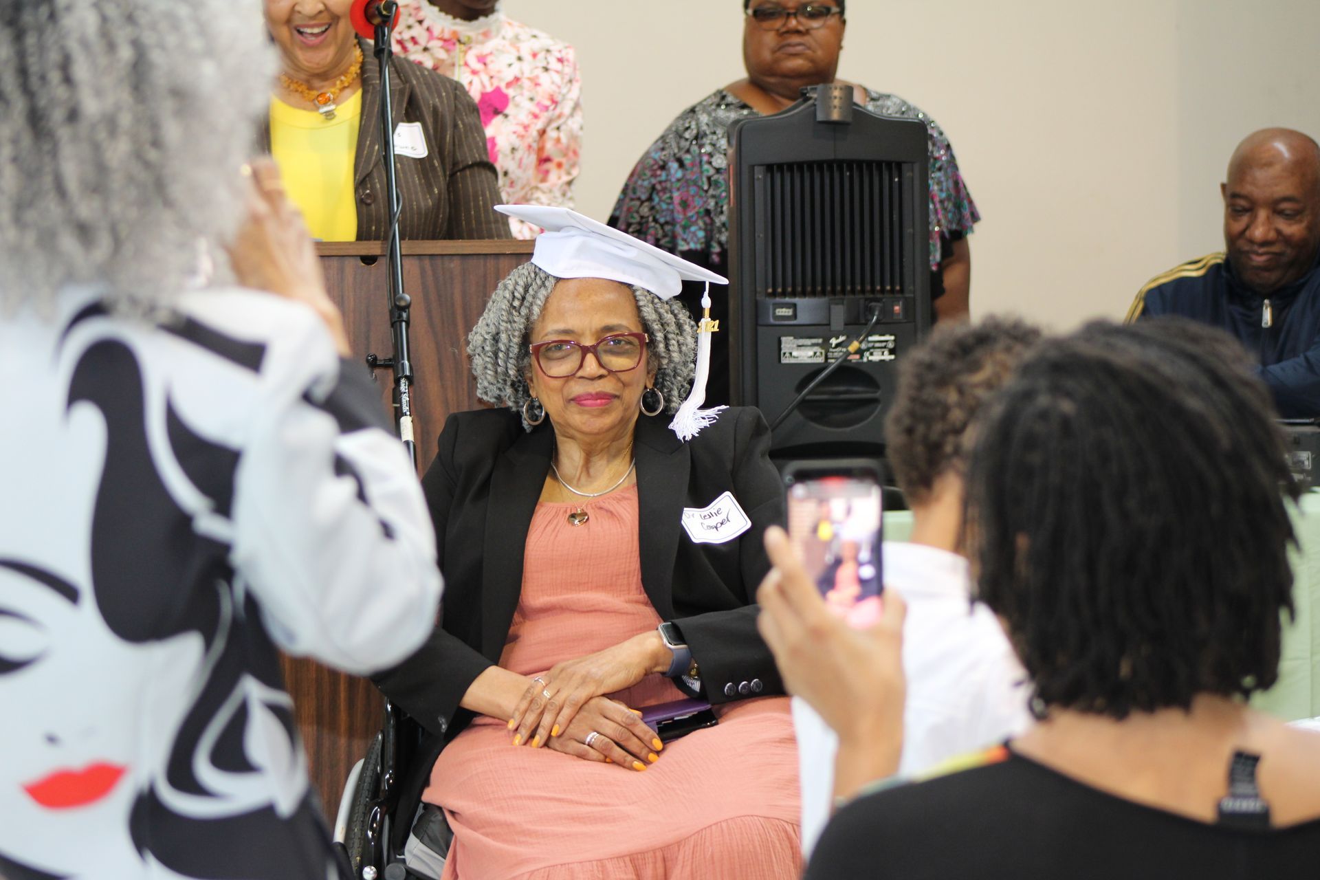 Woman in wheelchair, wearing graduation cap, being photographed by people at a celebration.