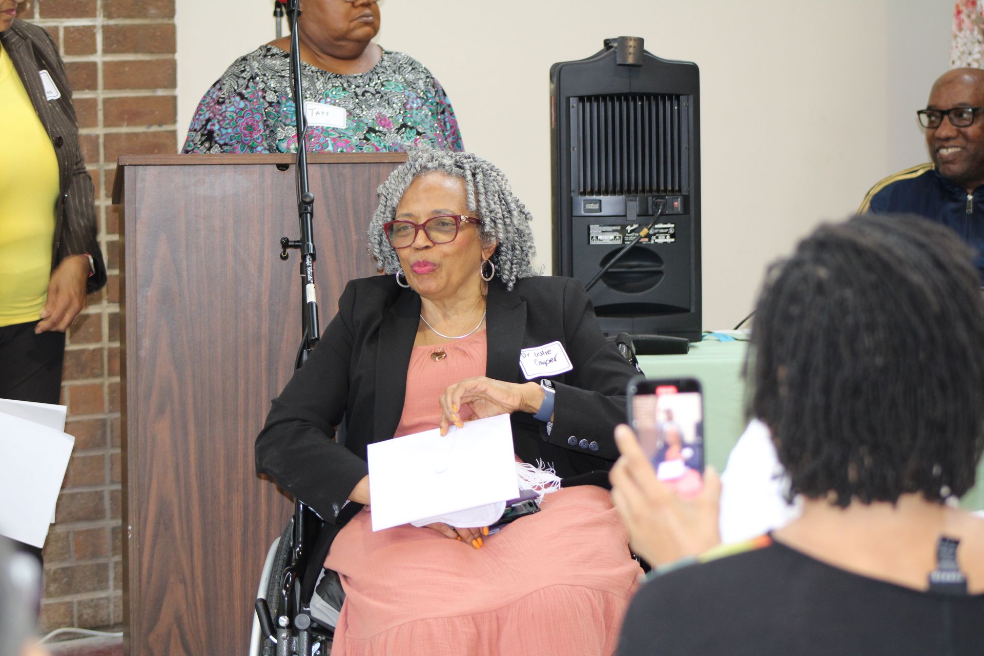 Woman in a wheelchair speaking, holding papers, at a podium. Others are nearby.