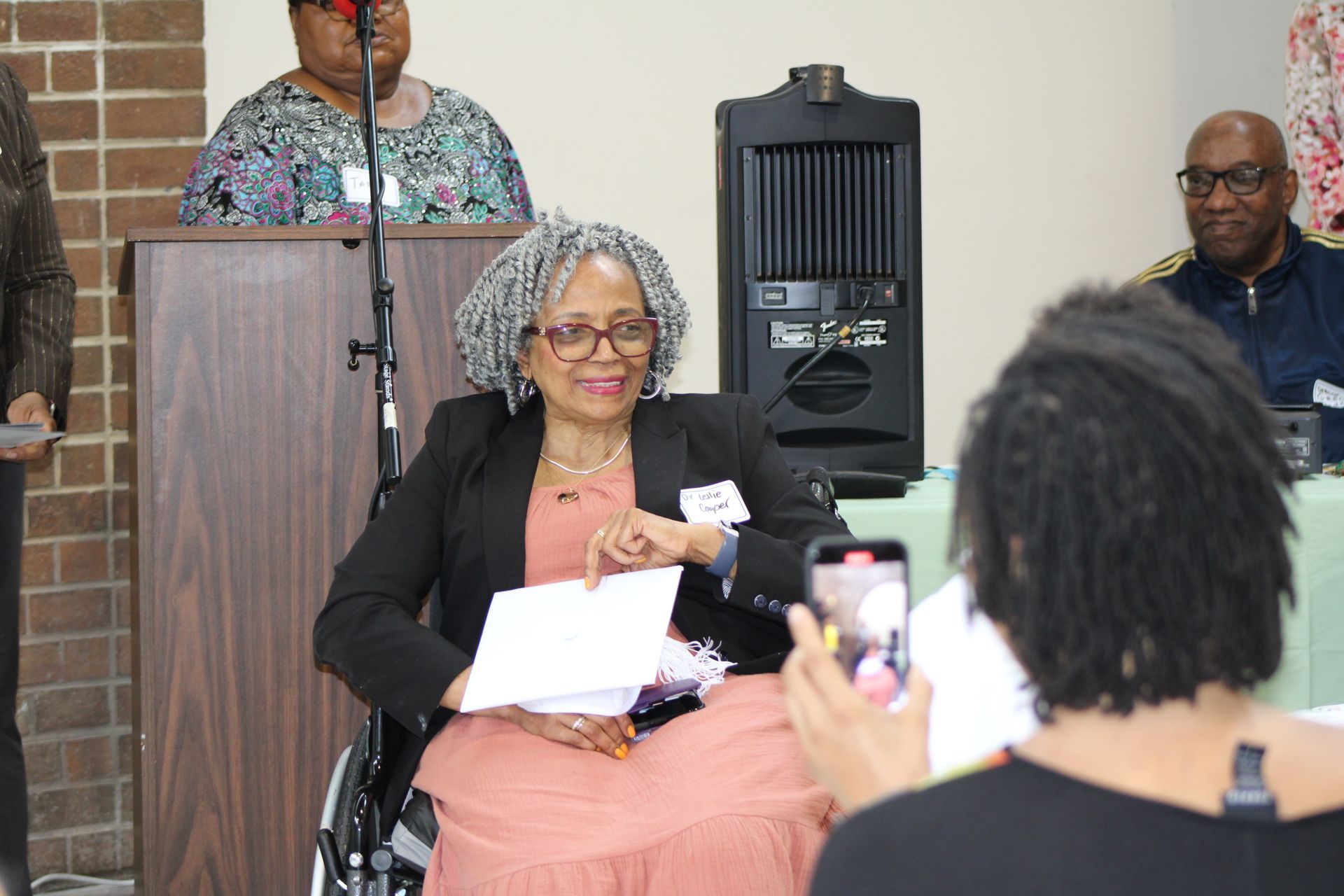 Woman in wheelchair smiles, holding papers, being photographed. Other speakers and podium in a meeting room.