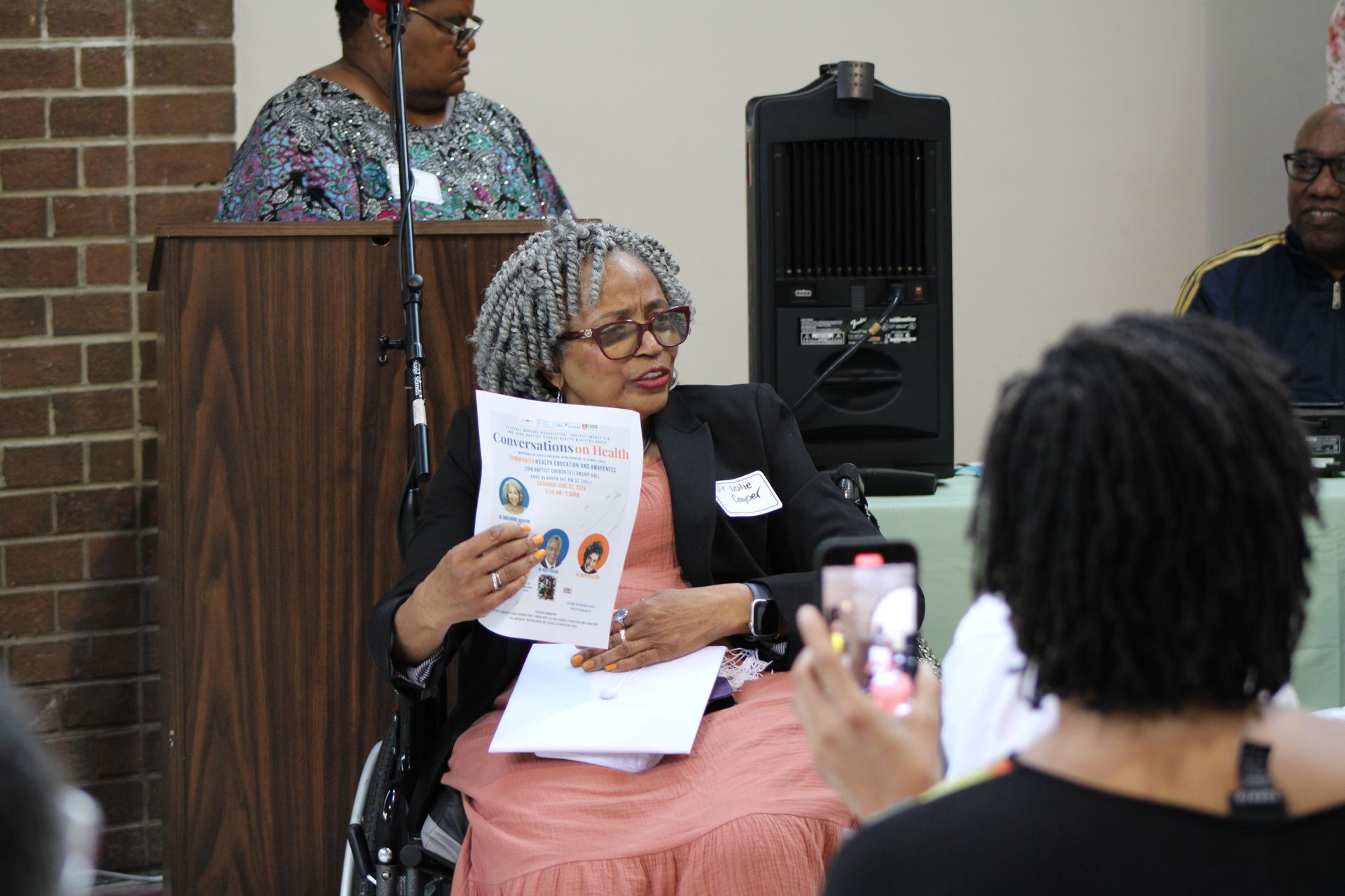 Woman in wheelchair speaking, holding paper with images, at an outdoor event.