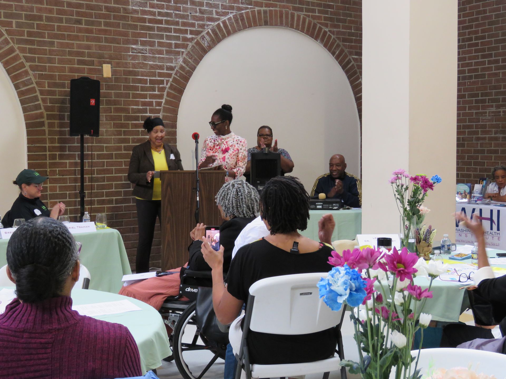 People at a meeting in a brick-walled room. Speakers at a podium, audience applauding, tables with flowers.