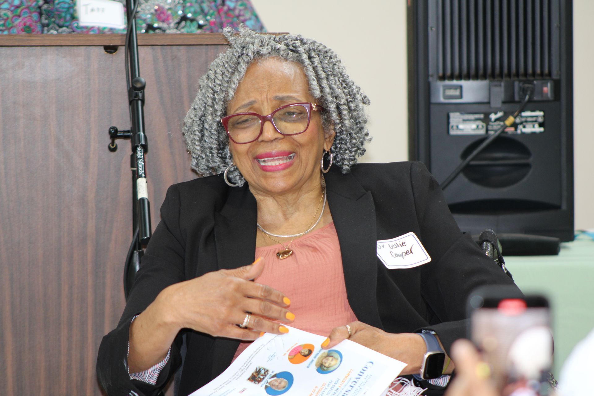 Woman in glasses and blazer smiles, gesturing while holding paper, speaker and microphone in background.