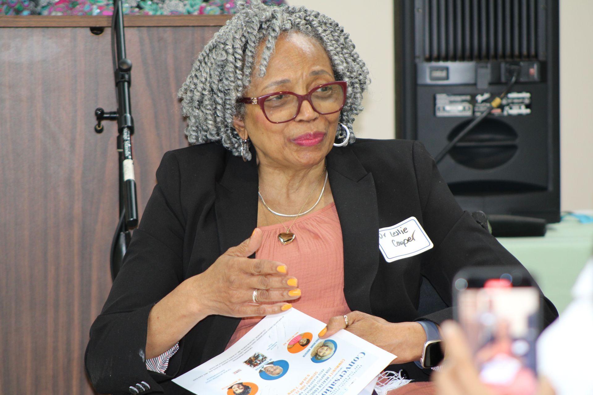Woman in glasses, black blazer, holding papers, speaking at an event with microphone and speaker.