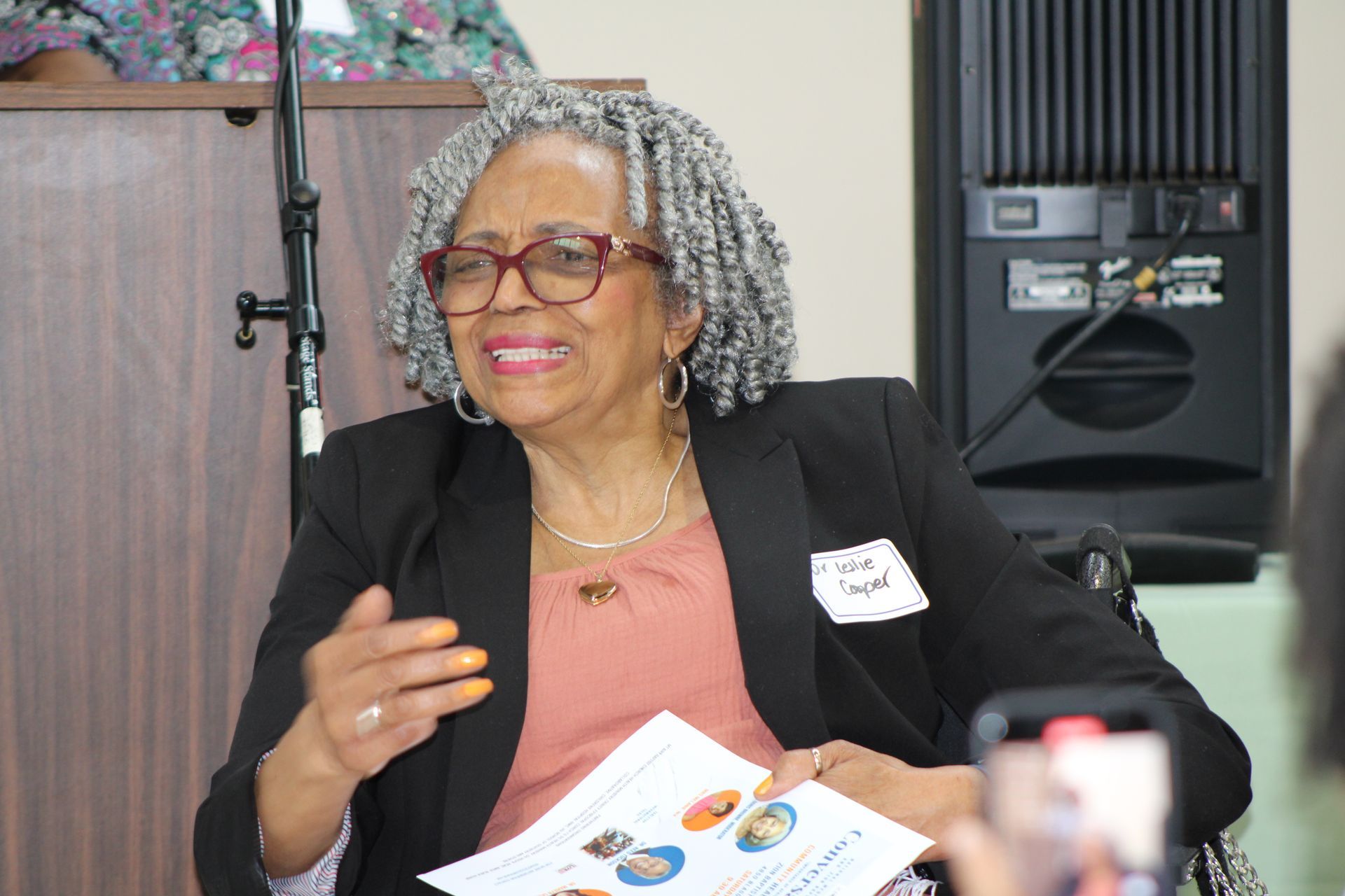 Woman with glasses smiles, gesturing during a presentation; holding papers; sitting near a speaker.