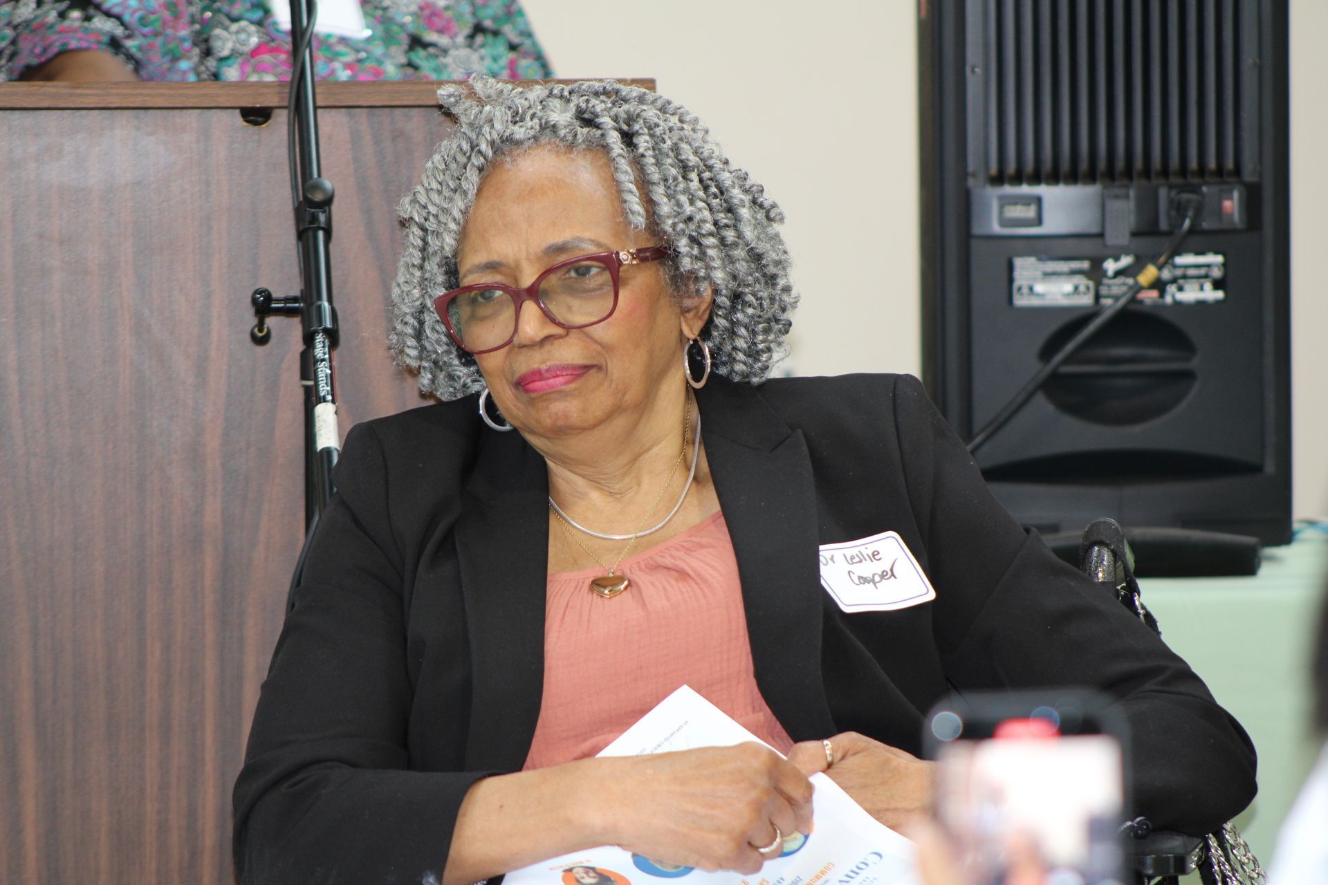 Woman in glasses and blazer at an event, holding papers.  She is in a wheelchair near a speaker.