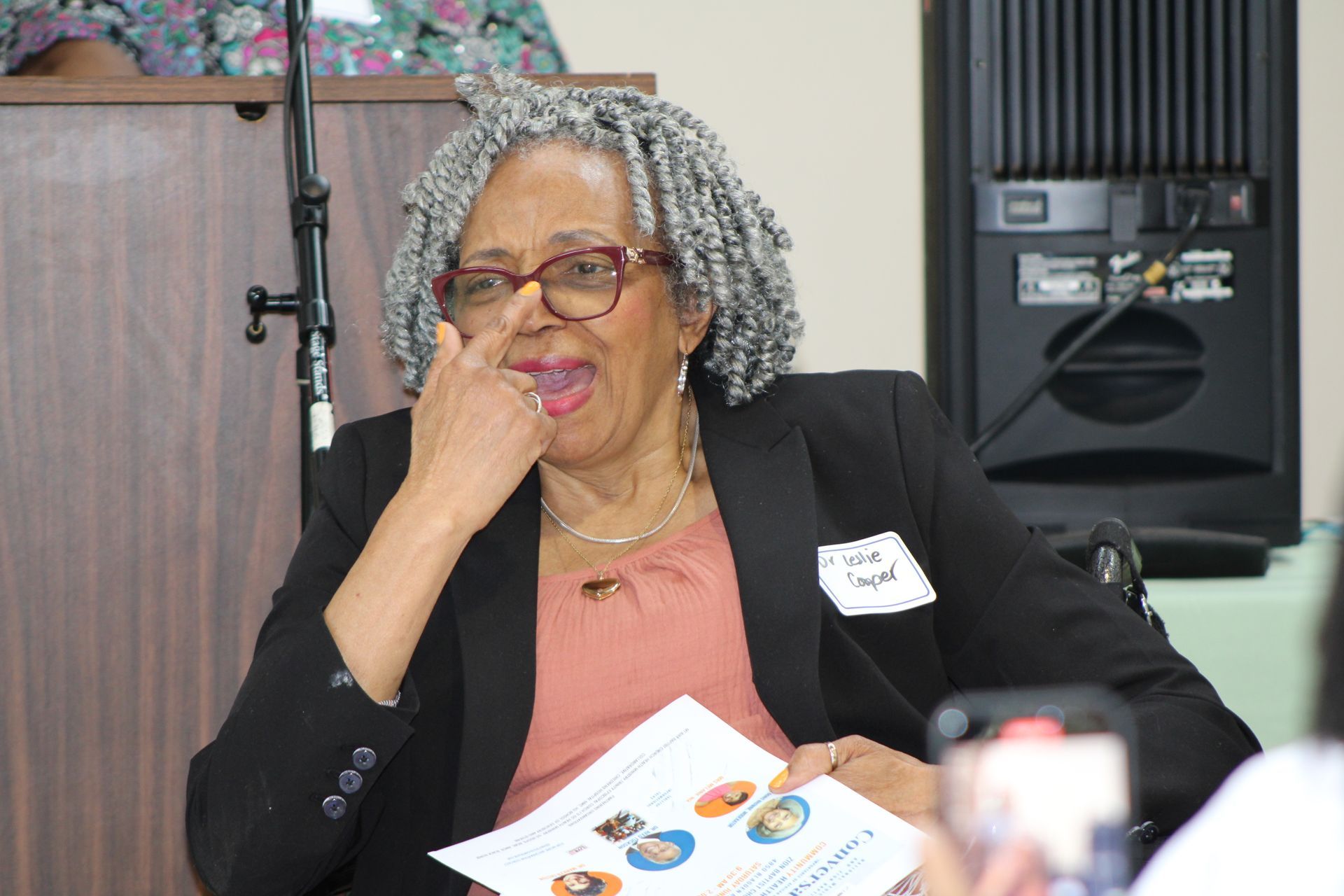 Woman in glasses points at her nose, holding a paper; she sits at a table near a speaker.