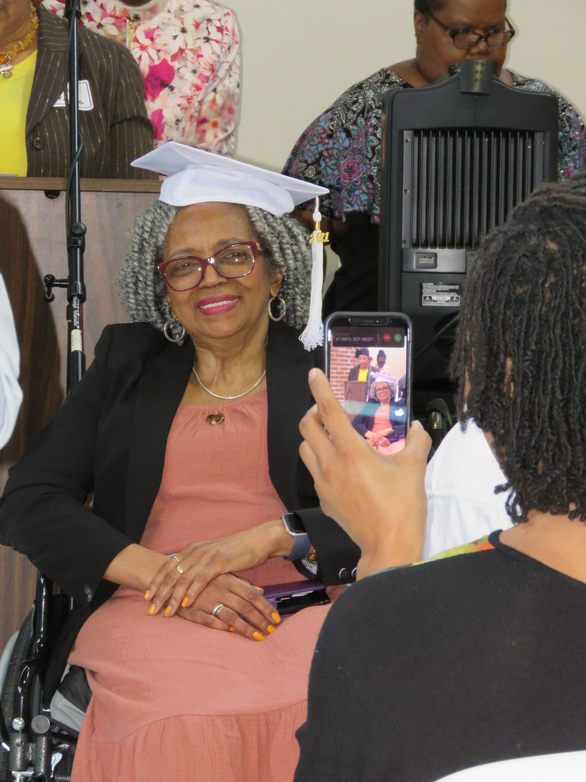 Woman in graduation cap smiles while being photographed, indoors. She wears a pink dress and black blazer.