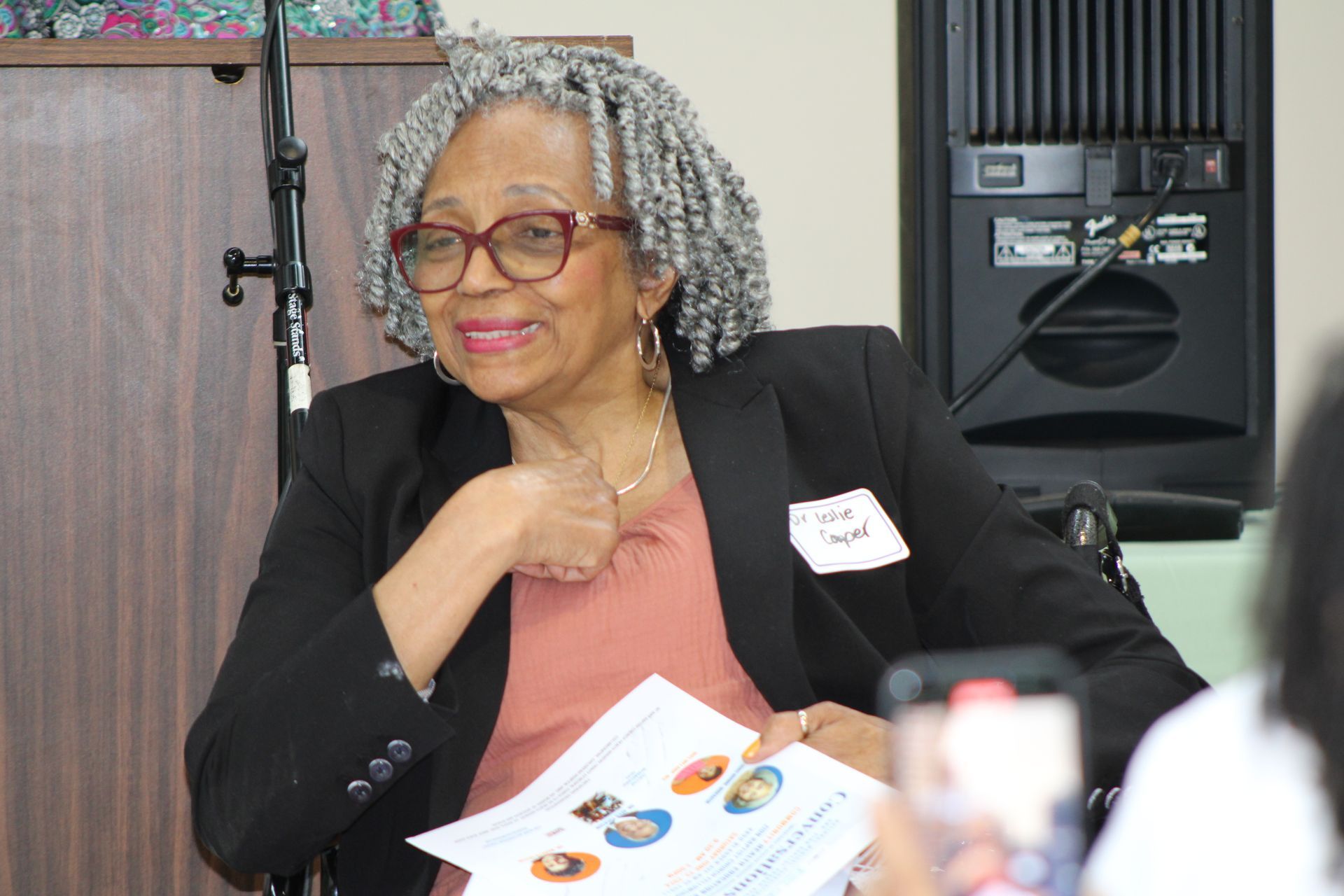Woman with gray hair and glasses in a black blazer smiling, holding papers at a presentation, speaking.