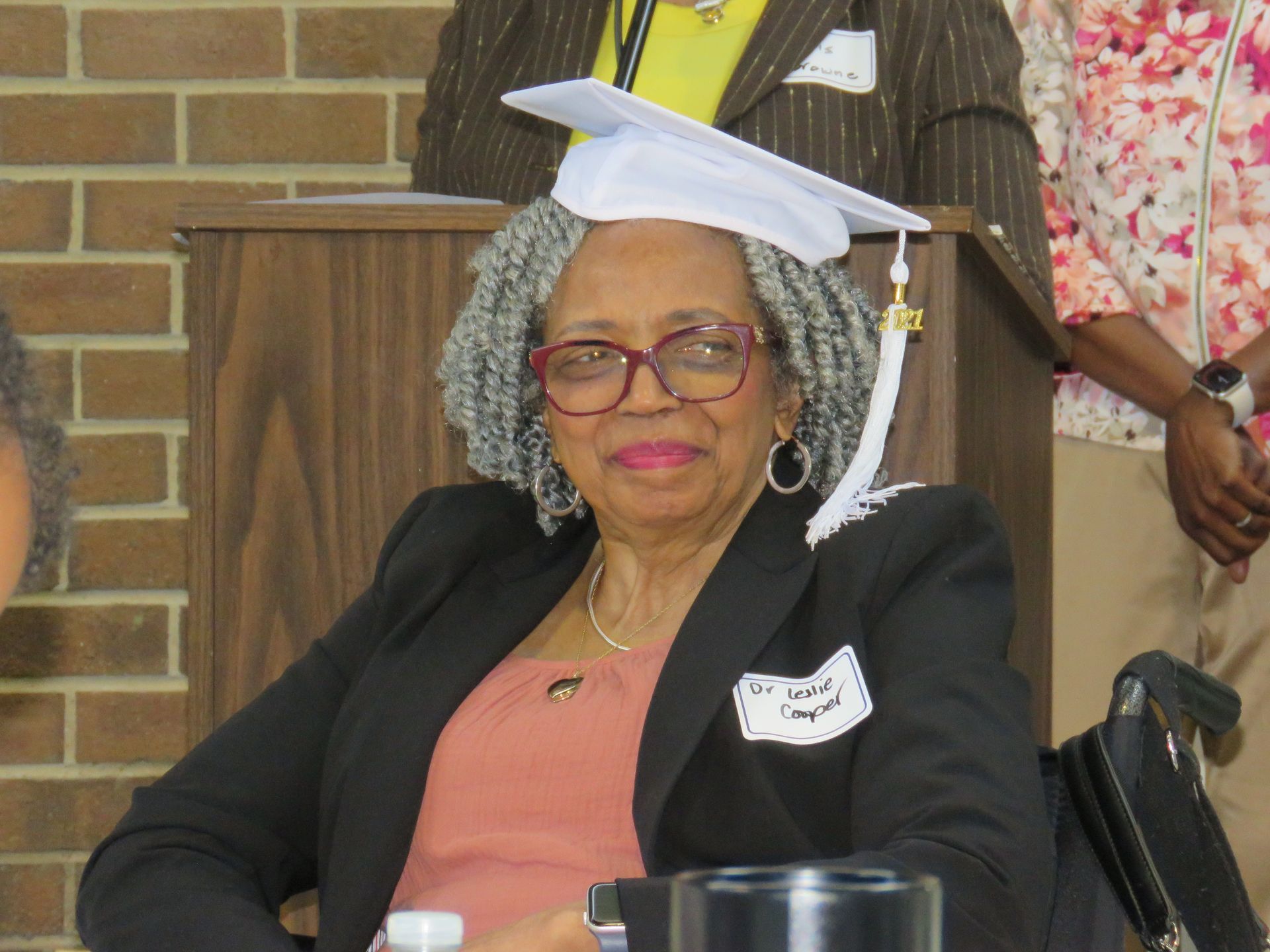 Woman in a black blazer and graduation cap smiles, seated near a podium.