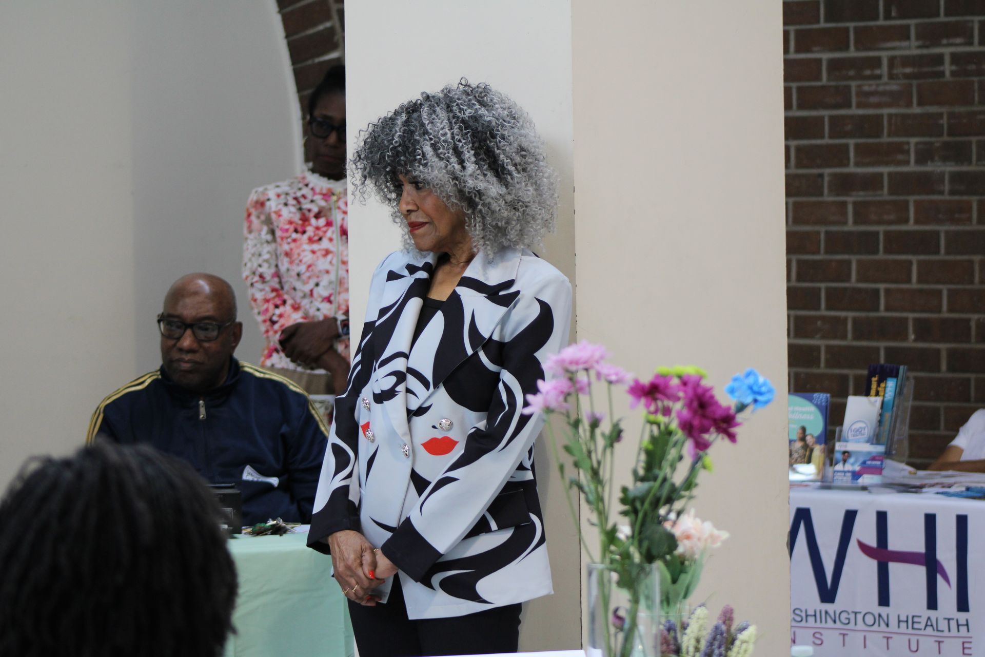 Woman in patterned jacket, speaking at event. Attendees and flowers are visible.