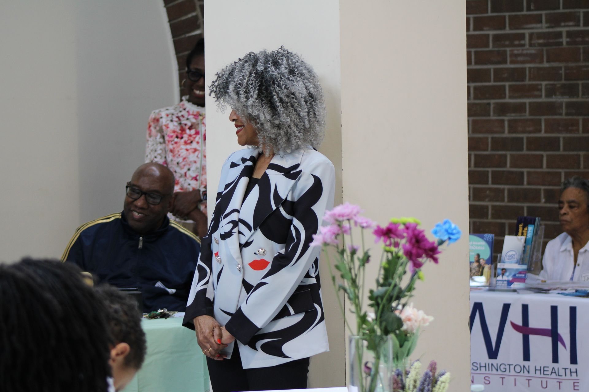 Woman with gray curly hair wearing a black and white jacket stands at an event. People and flowers are in view.