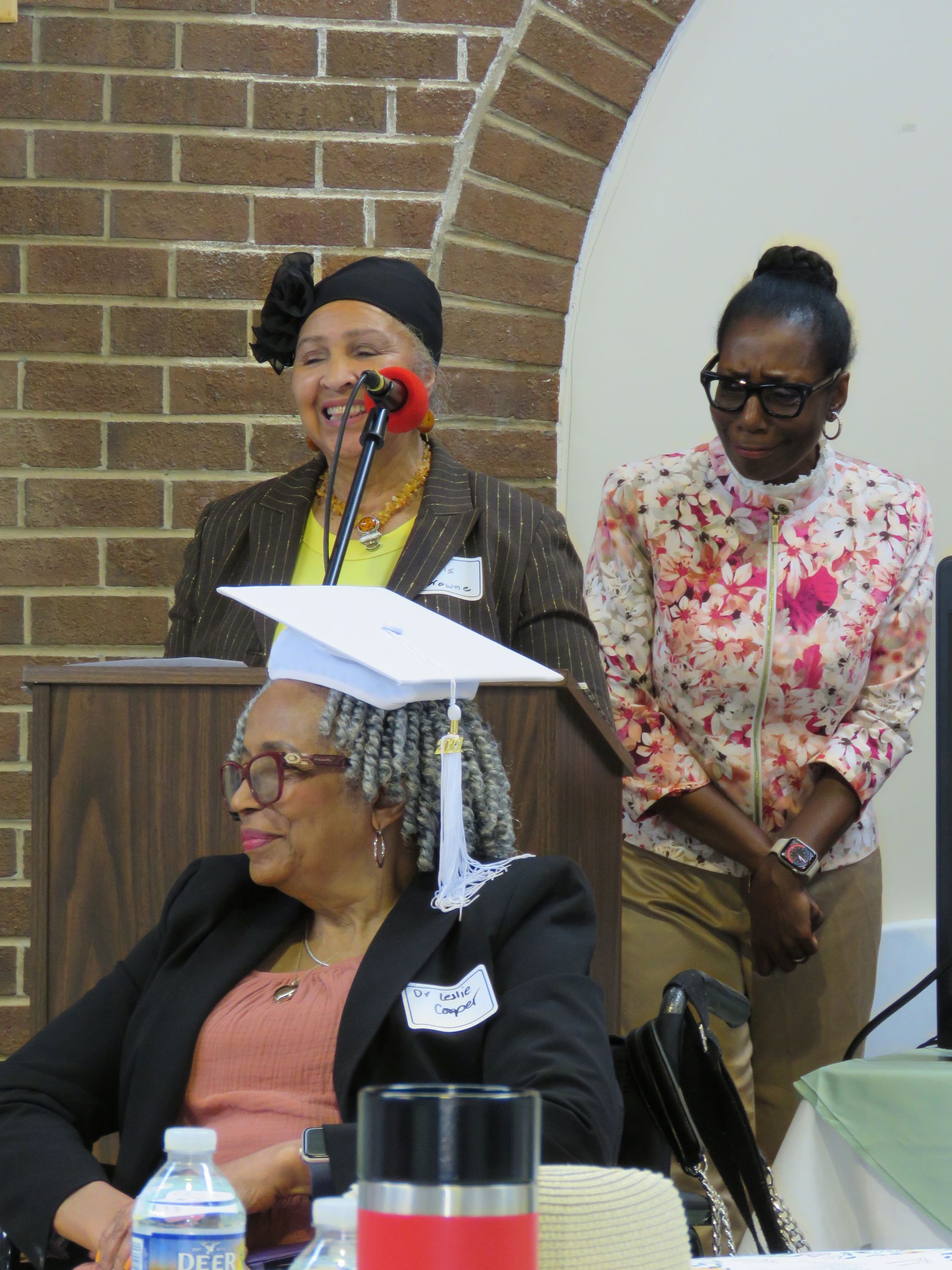 Three women at a podium: One speaking, two watching. Brick wall background, indoors. One has a mortarboard.
