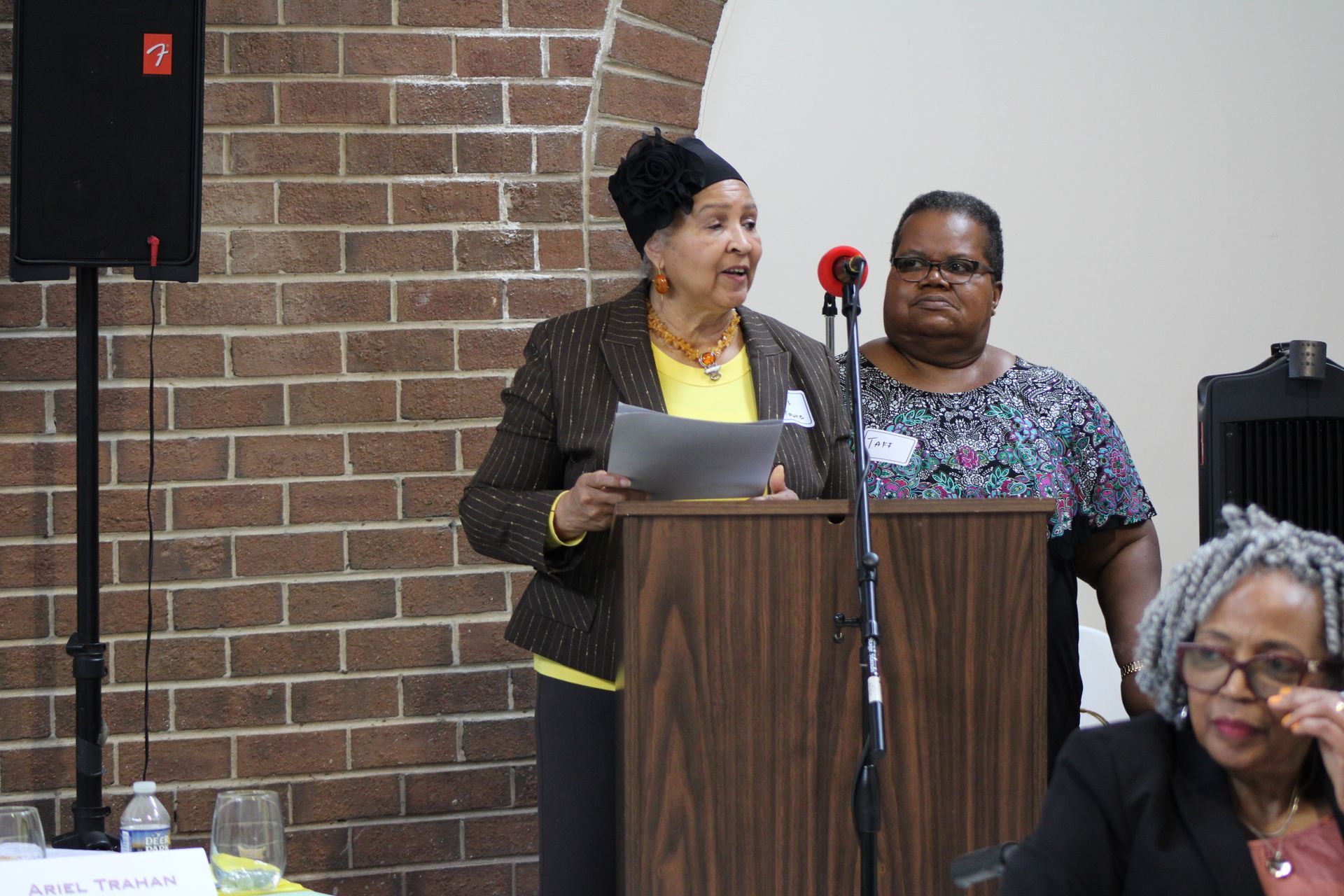 Woman speaking at a podium, another woman stands behind her. A third woman sits at a table. Brick wall background.