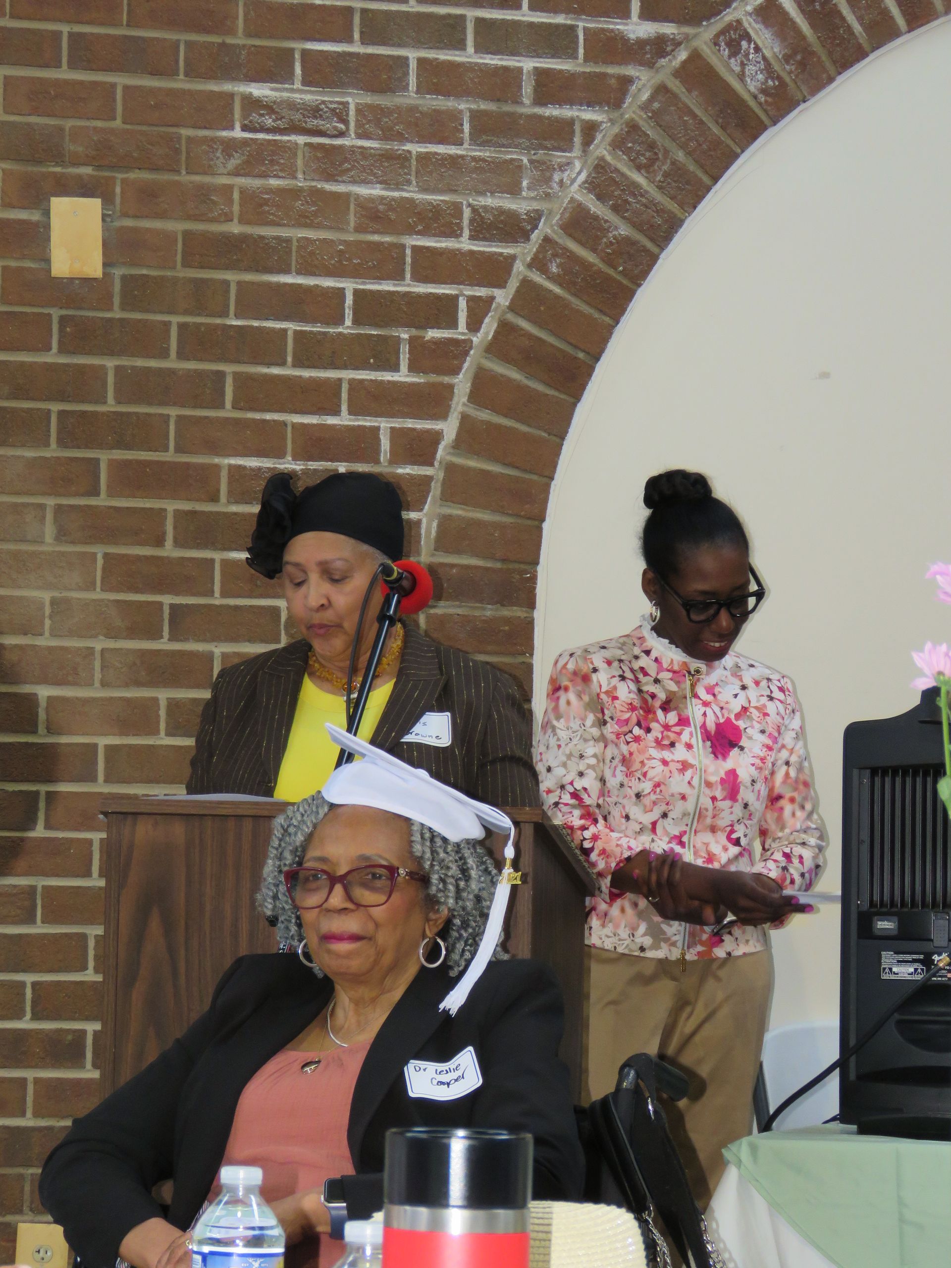 Three women at a podium and table. One in a graduation cap, others wearing hats and speaking, a brick arch in the background.